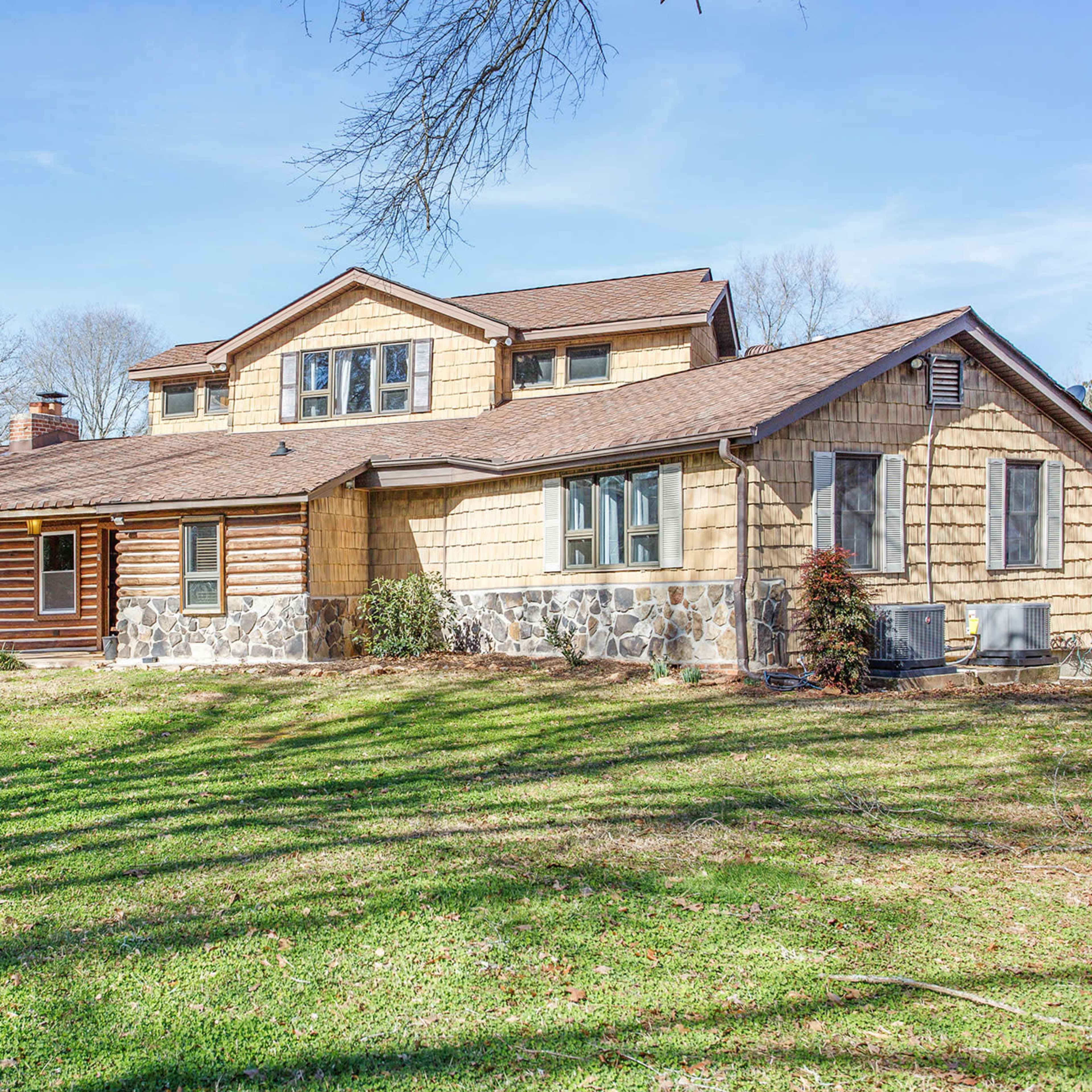 A two-story house with a combination of wooden and stone siding is situated on a grassy lawn, surrounded by trees.
