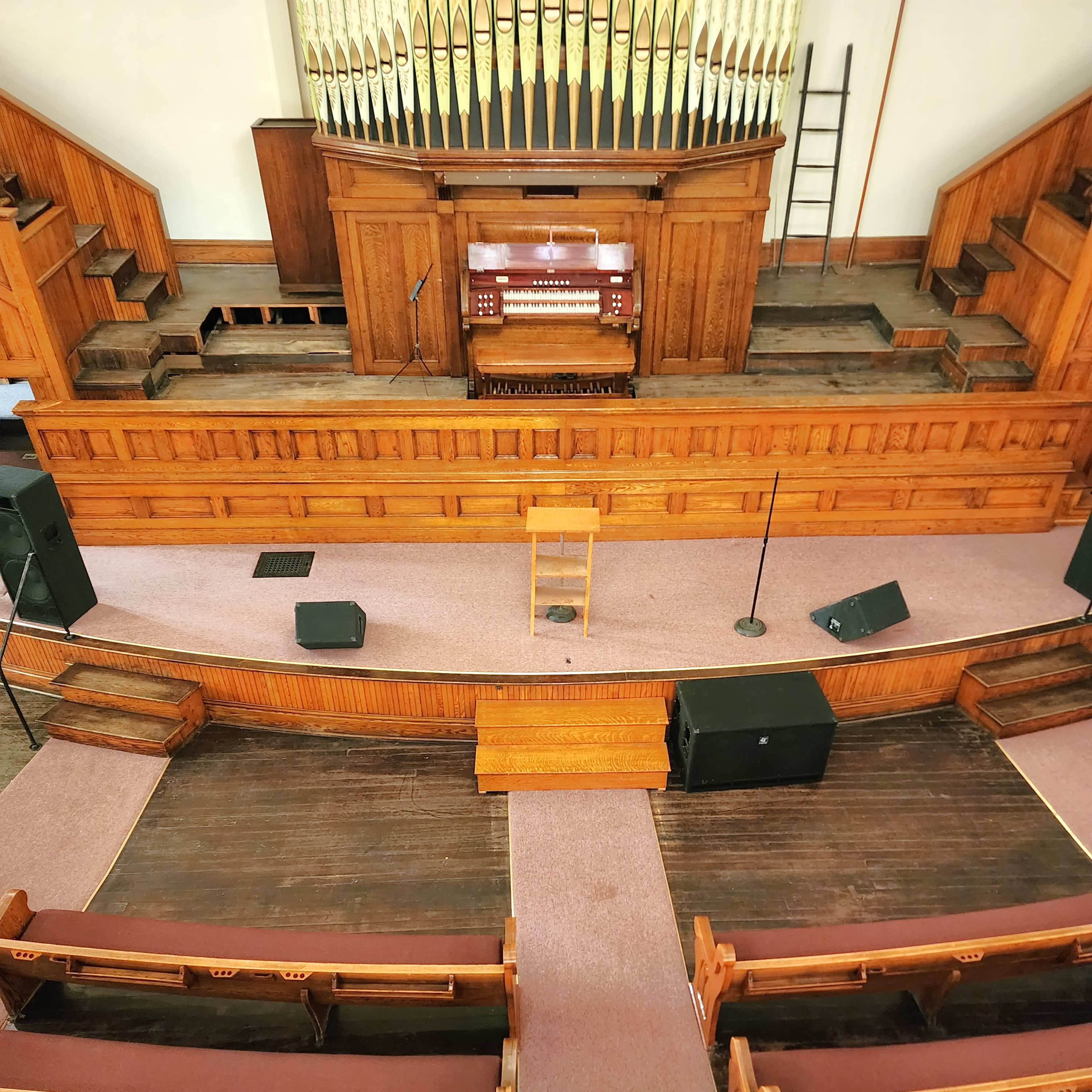 A wooden auditorium with a stage featuring an organ, seating rows arranged in front, and audio equipment positioned on the floor.