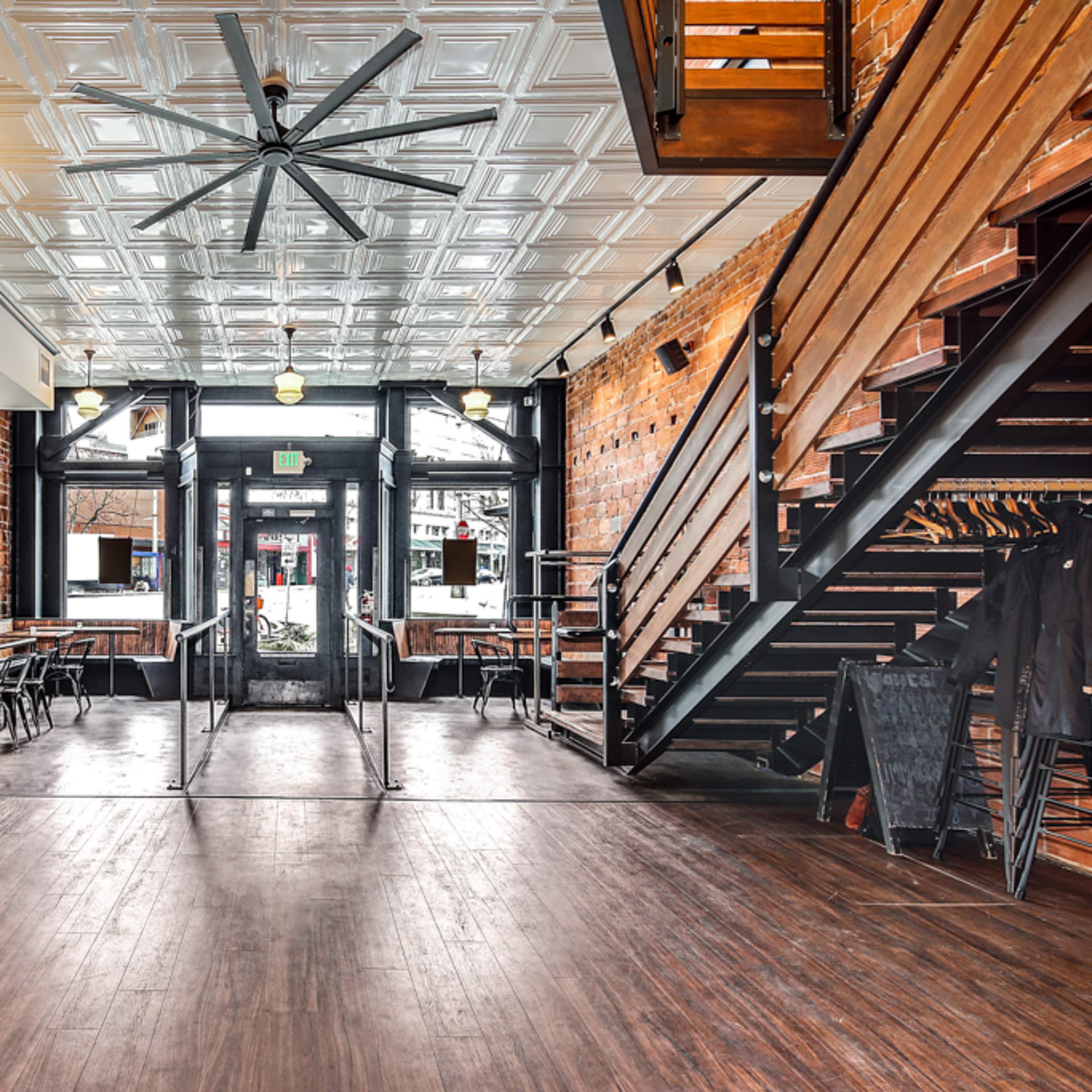 A spacious interior of a restaurant featuring exposed brick walls, wooden floors, and a large ceiling fan, with tables and a staircase visible.