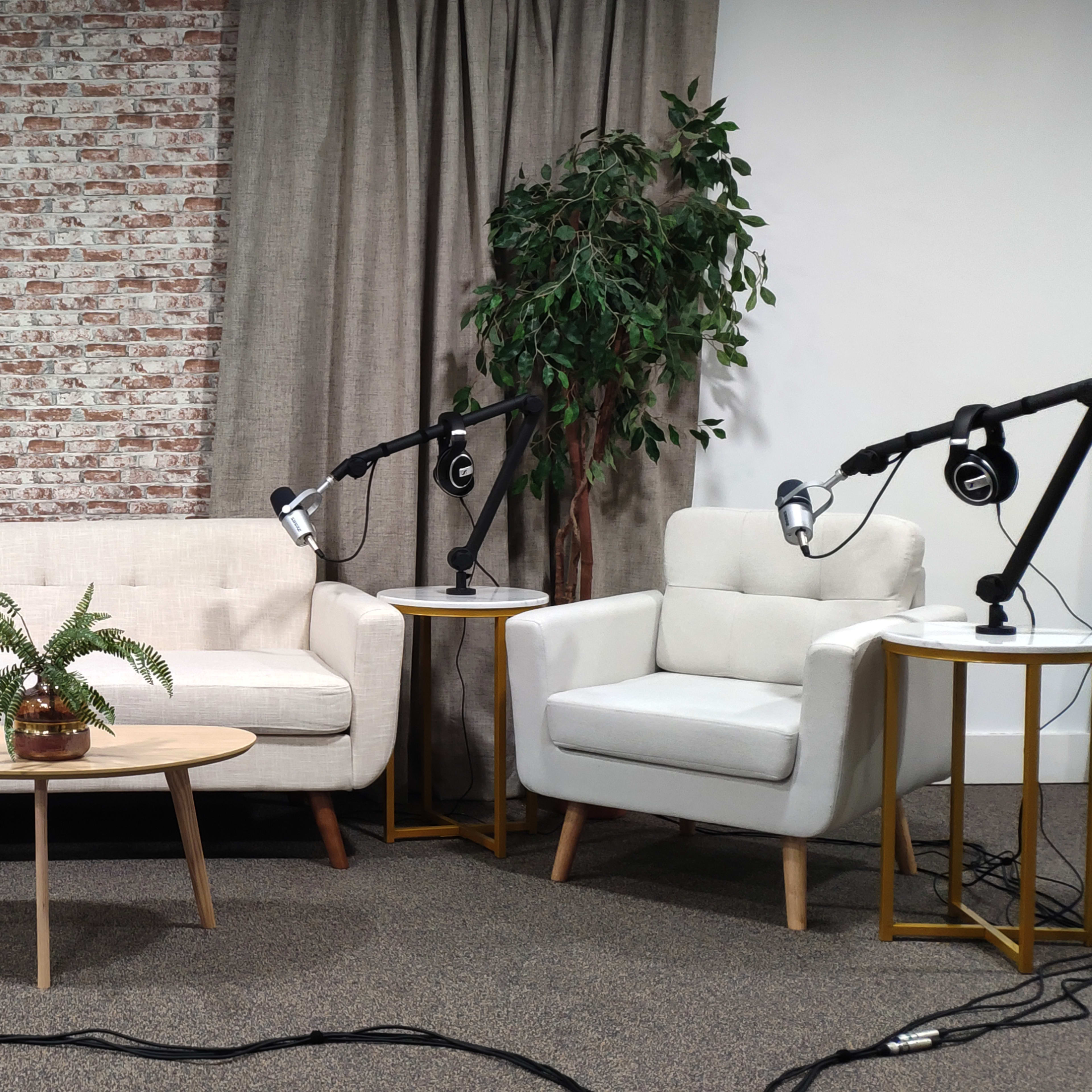 A small, professionally arranged interview or talk show set featuring a white sofa, two armchairs, potted plants, and microphone stands on either side.