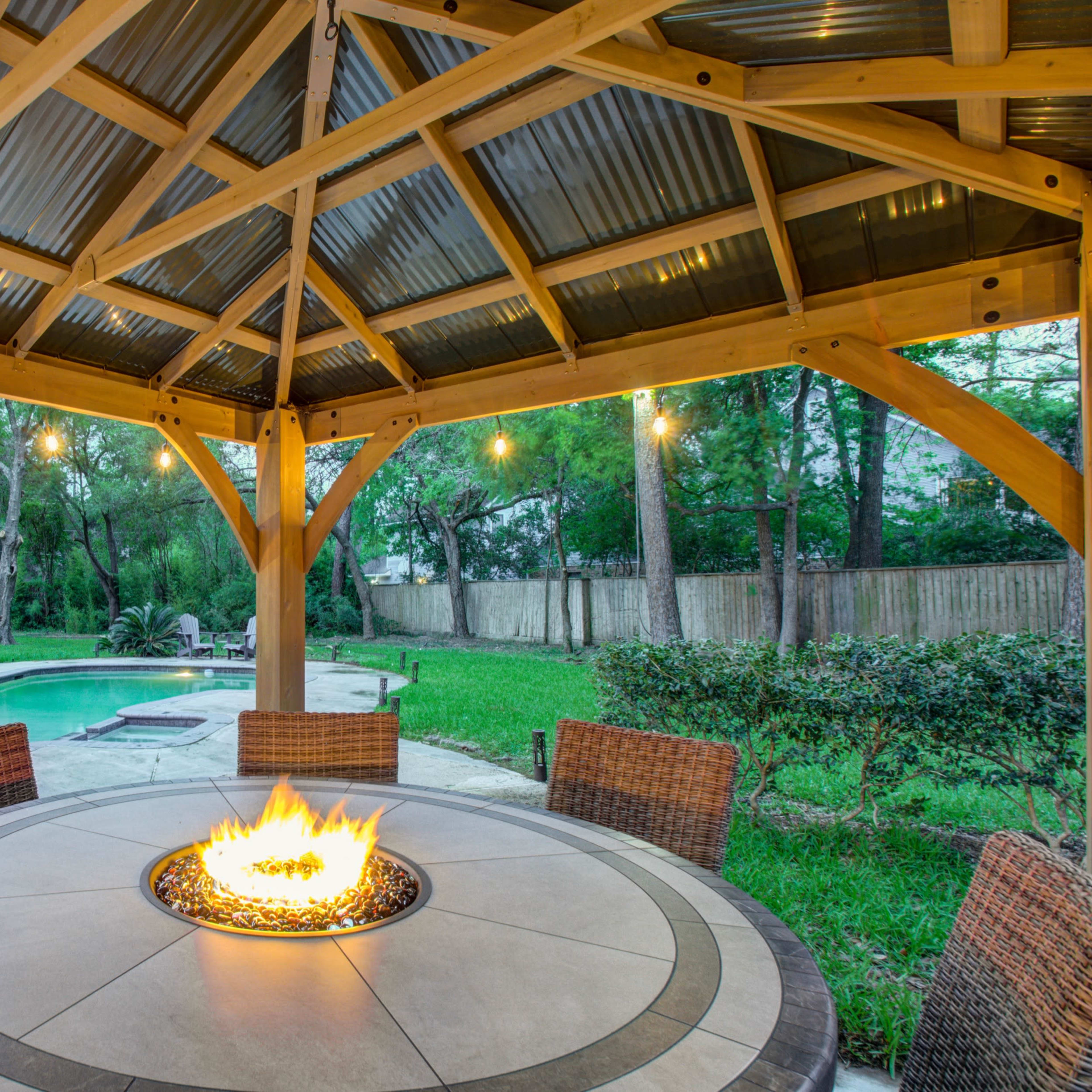 A gazebo with a fire pit is situated beside a swimming pool and surrounded by green grass and trees.