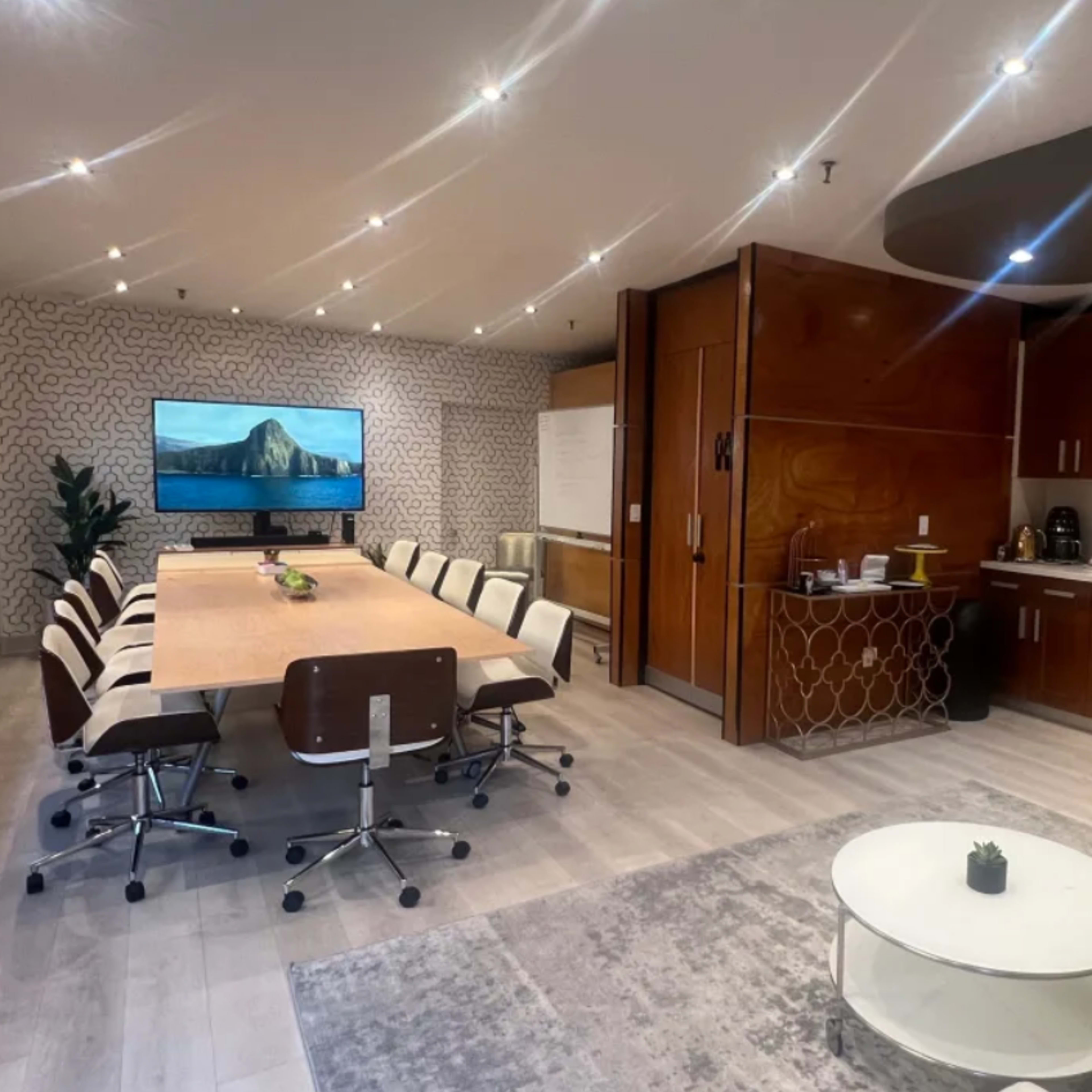 A modern conference room with a large table surrounded by ergonomic chairs, a wall-mounted TV, and a kitchenette area.