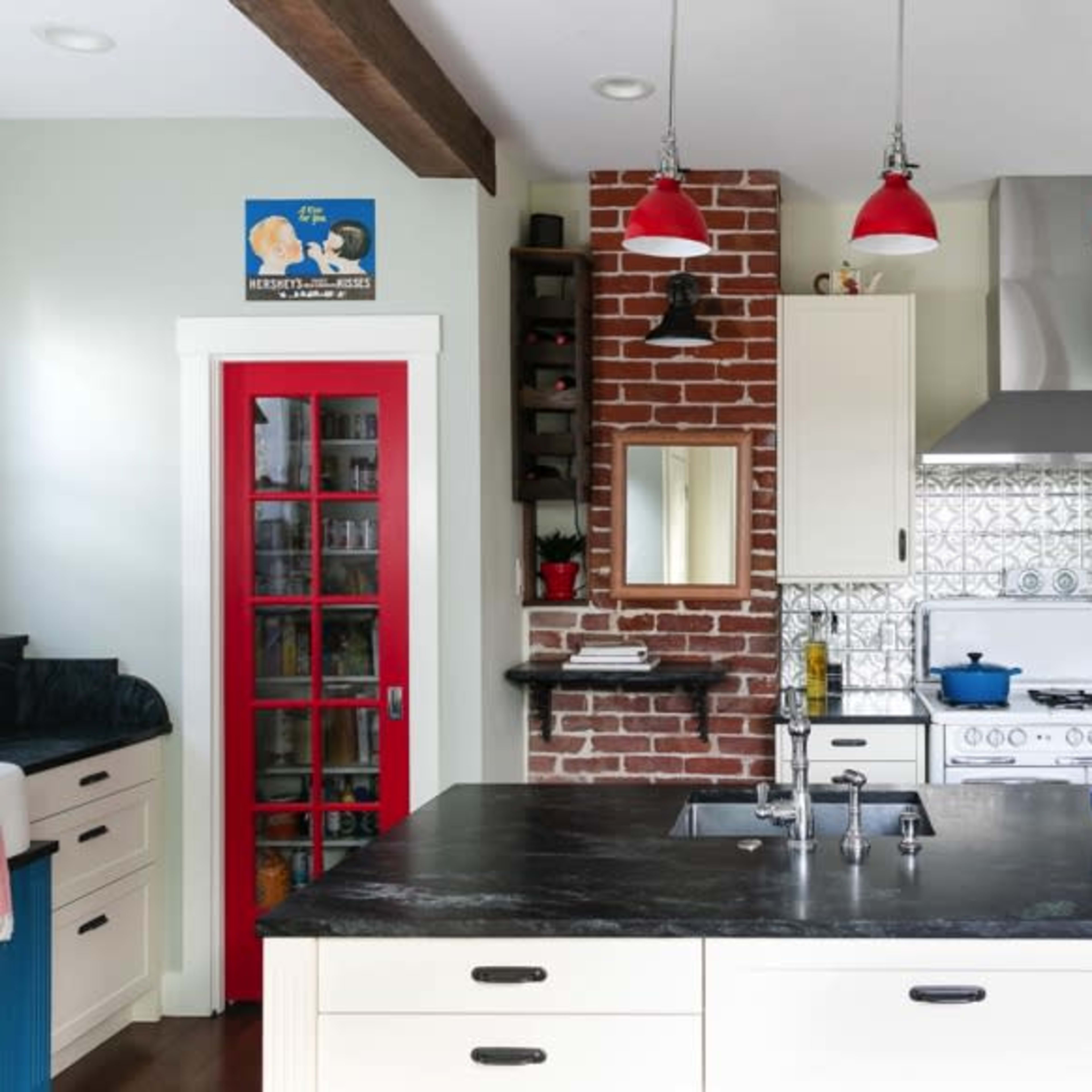 A modern kitchen features a center island with a black countertop, white cabinets, a red accent door, metal backsplash, and red pendant lights hanging from exposed beams.
