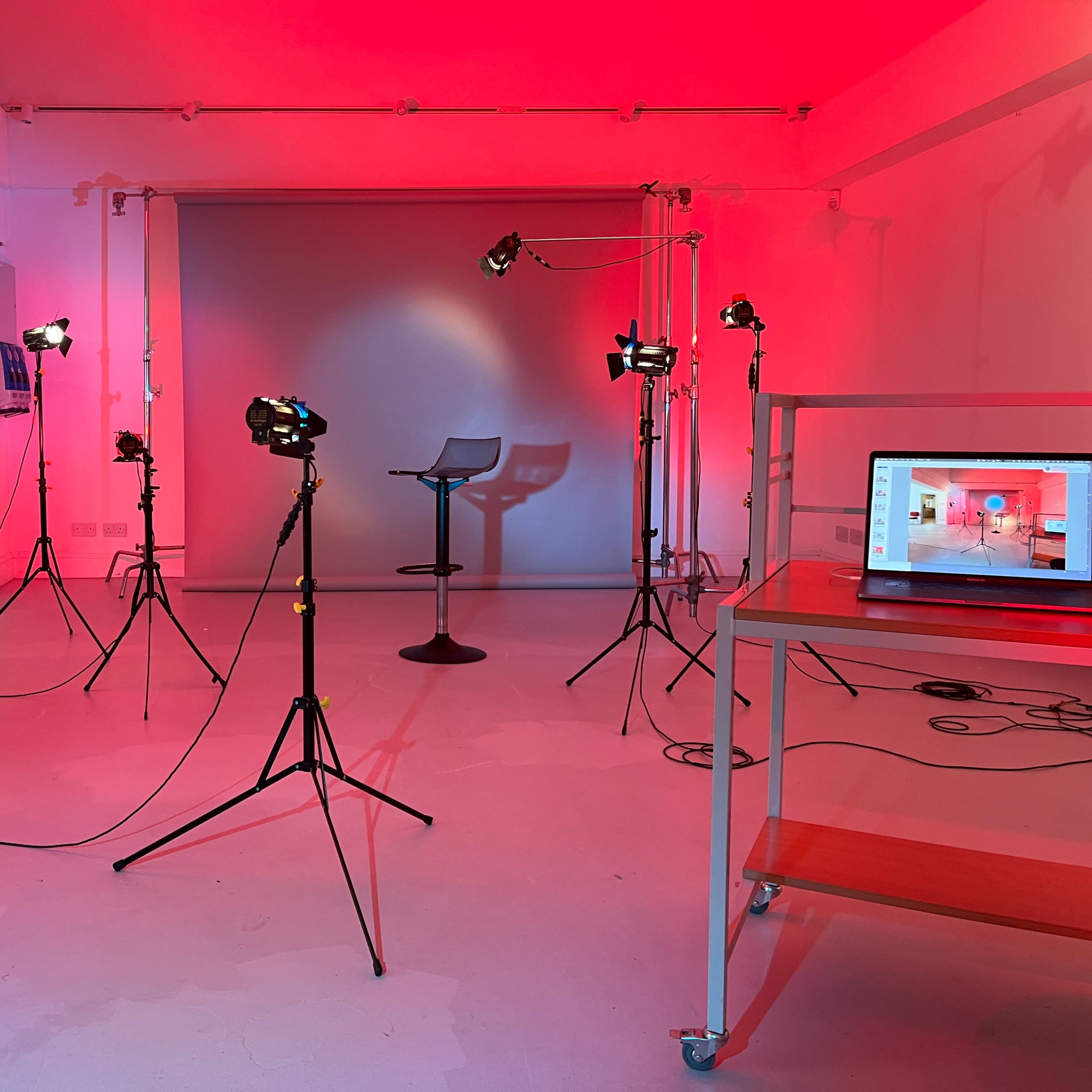 A studio space with red lighting, equipped with various photo lights on tripods, a stool, and a laptop on a table.