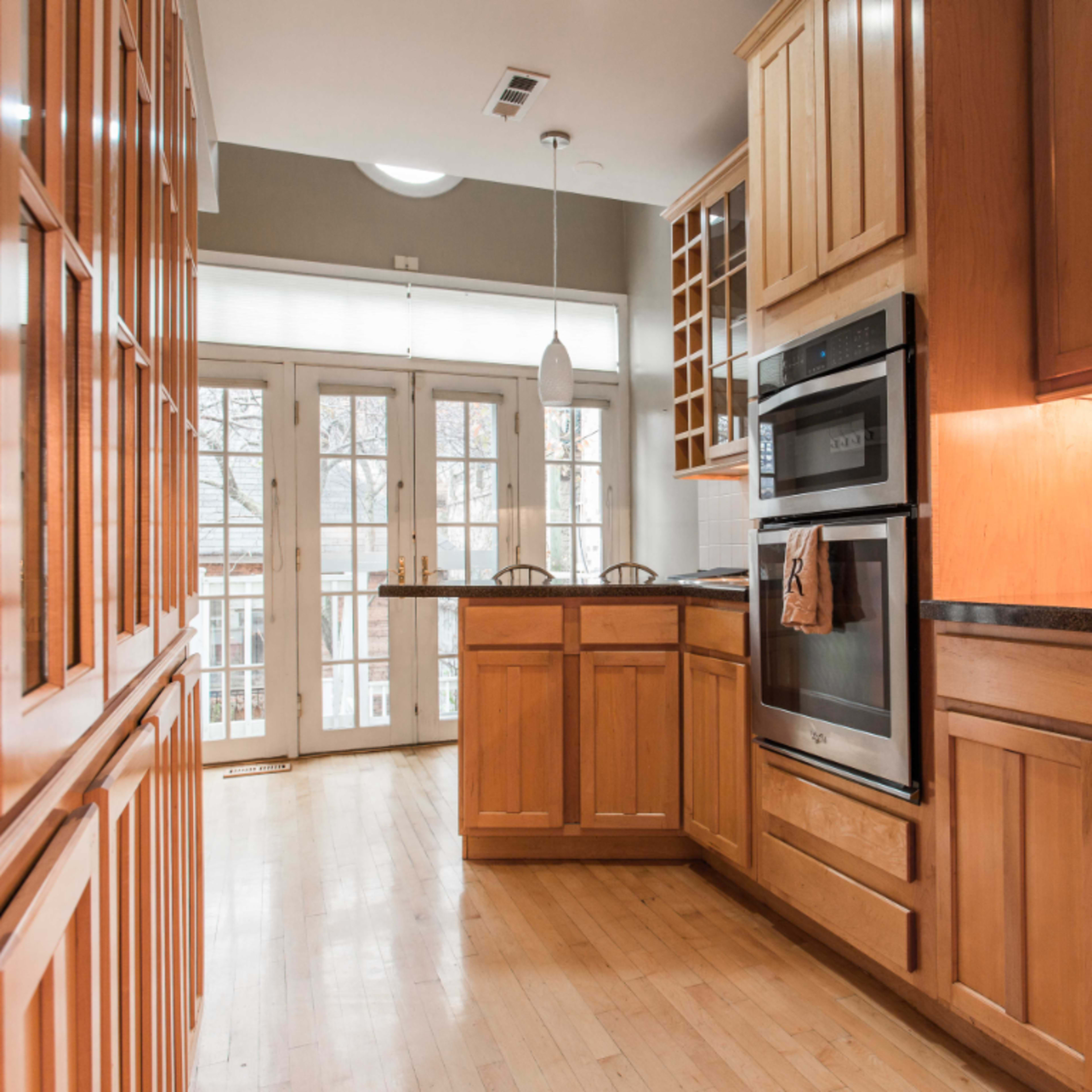 A narrow kitchen features wooden cabinets, a double oven, and a view of a patio through glass doors at the end of the room.