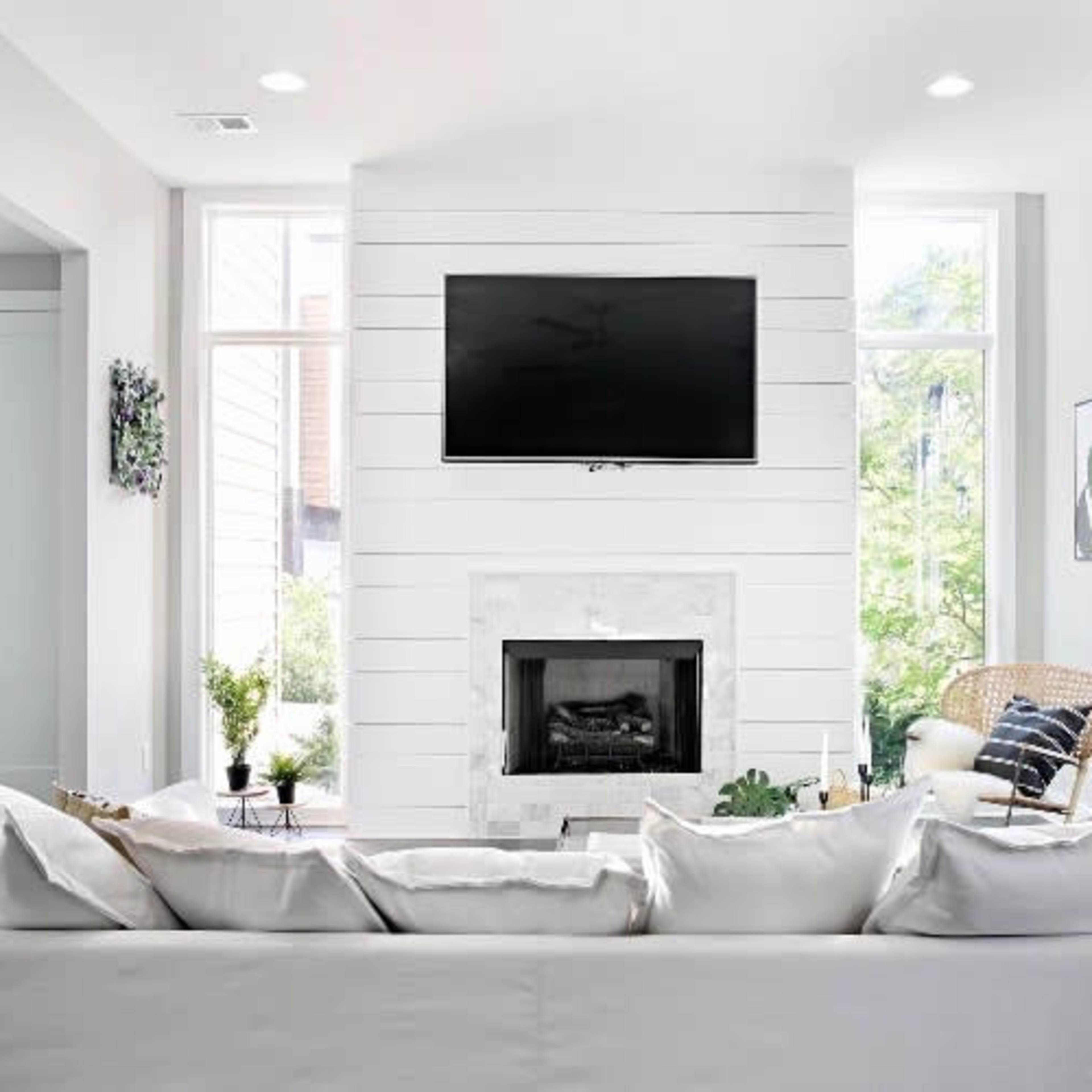A modern living room features a wall-mounted television above a marble-accented fireplace, with large windows flanking the space and a neutral color palette.