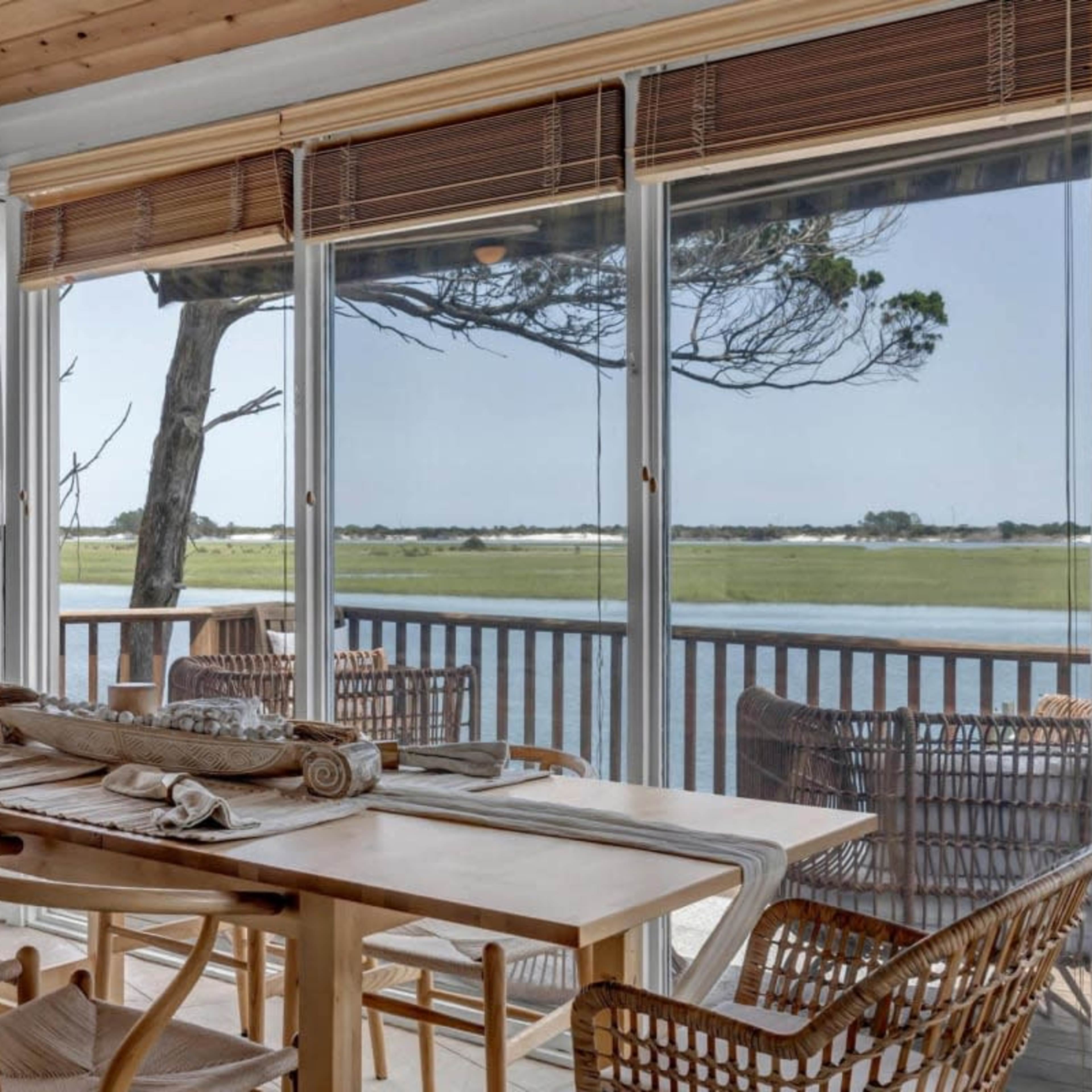 A dining area features a wooden table set with linens and overlooks a peaceful view of a river through large glass windows, framed by wooden beams and wicker furniture.