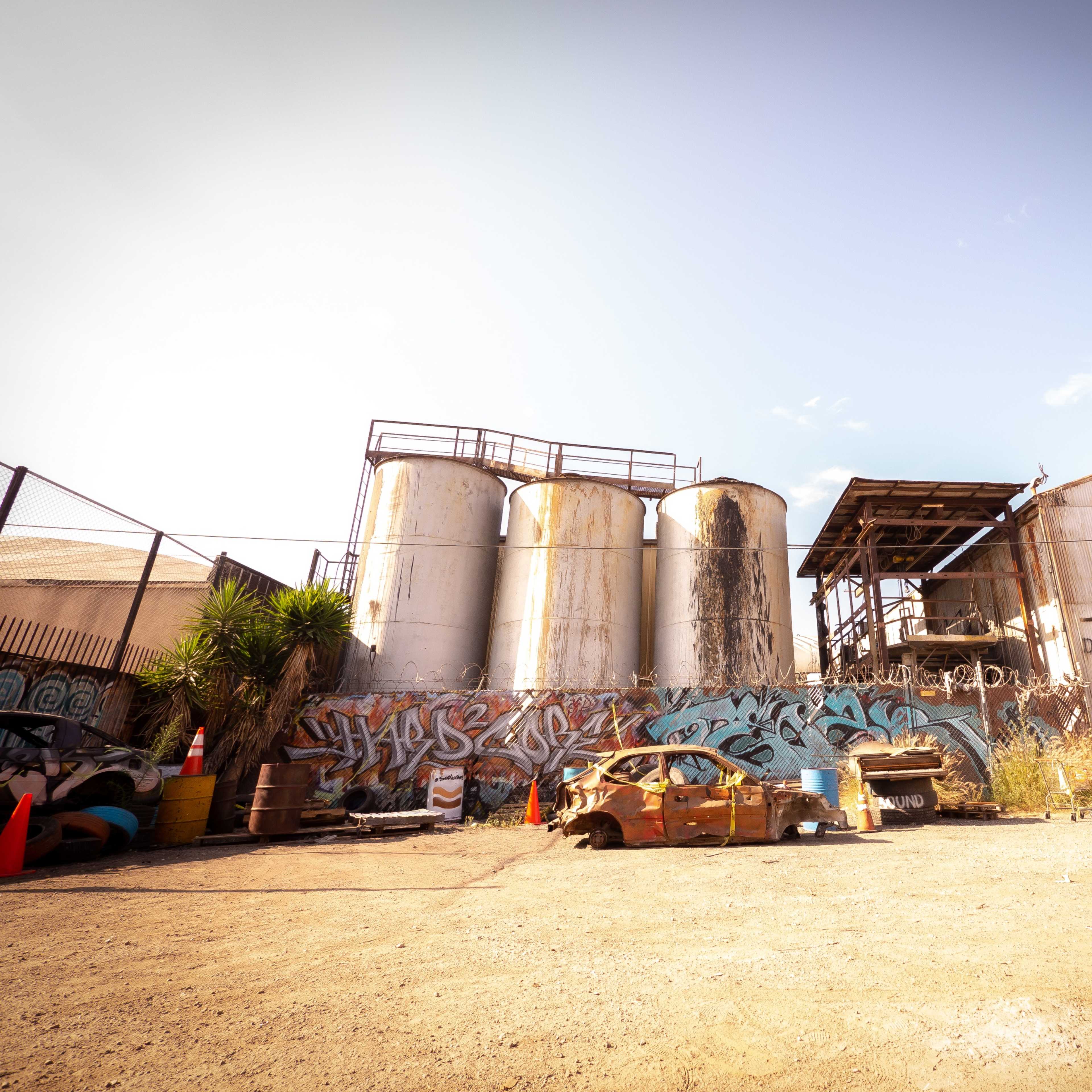 A deserted industrial area with three large silos, a dilapidated building, and a rusted car covered in graffiti.