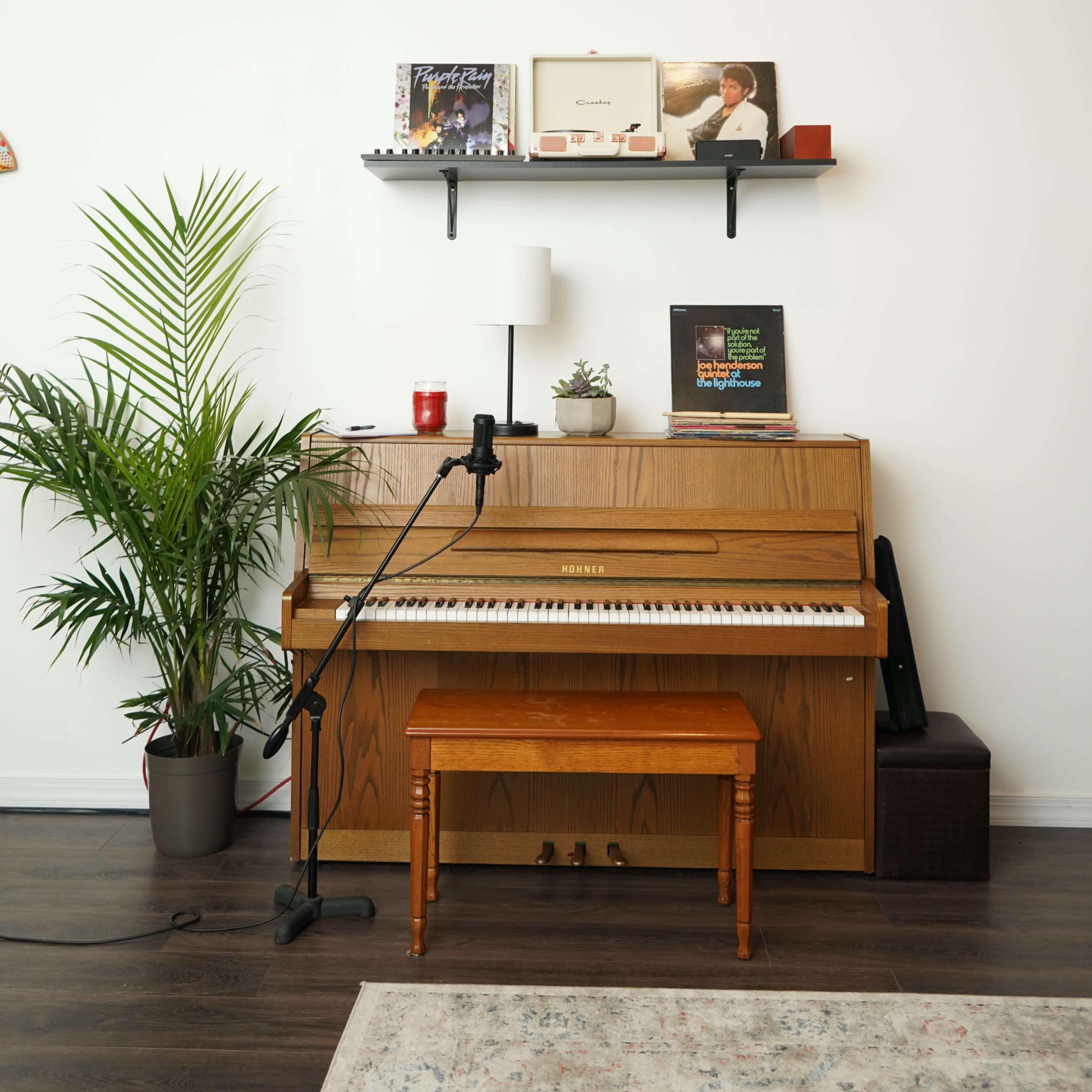A wooden piano with a matching bench is positioned beside a plant, under a shelf holding records and décor items.