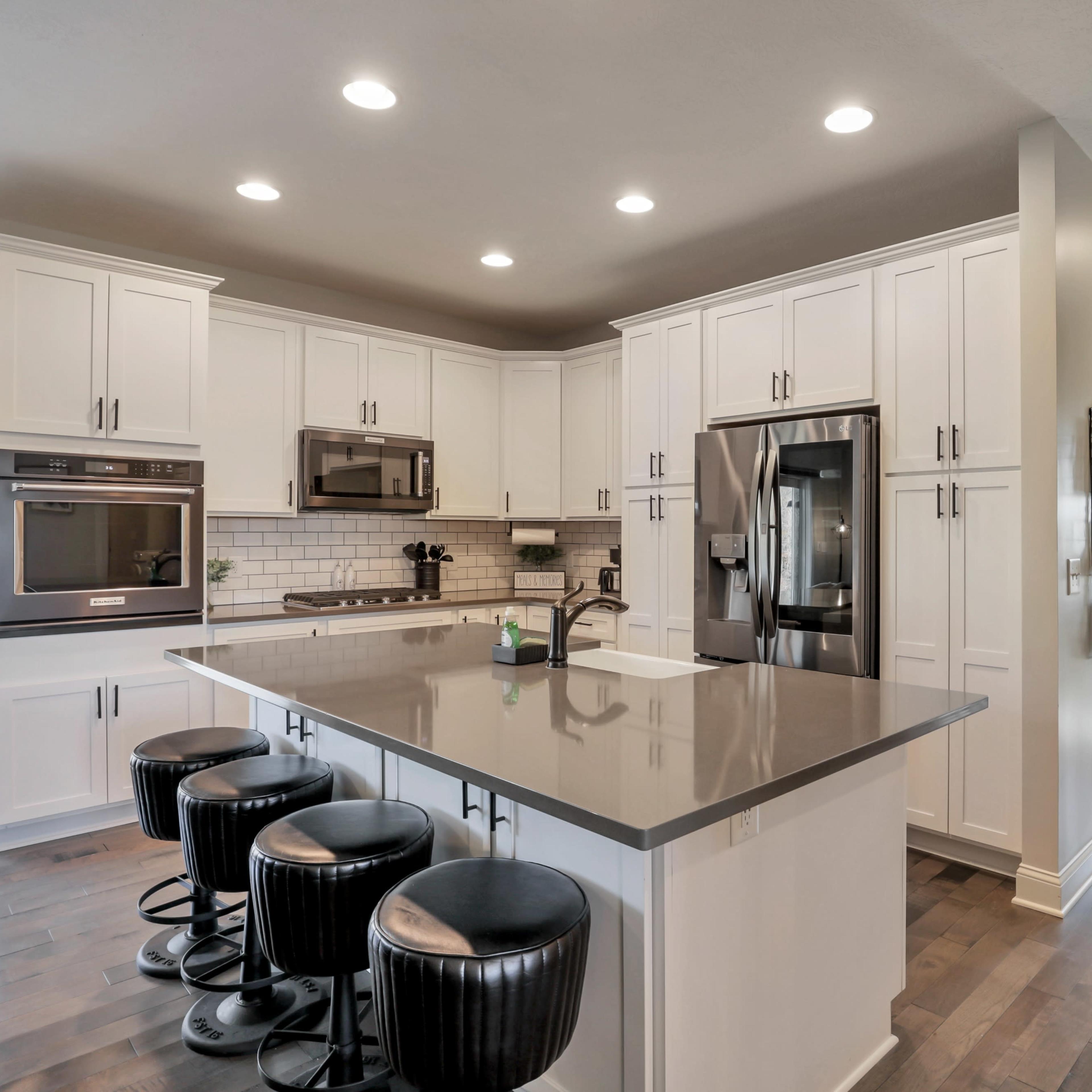 A modern kitchen with white cabinetry, stainless steel appliances, a spacious island with four black bar stools, and hardwood flooring.