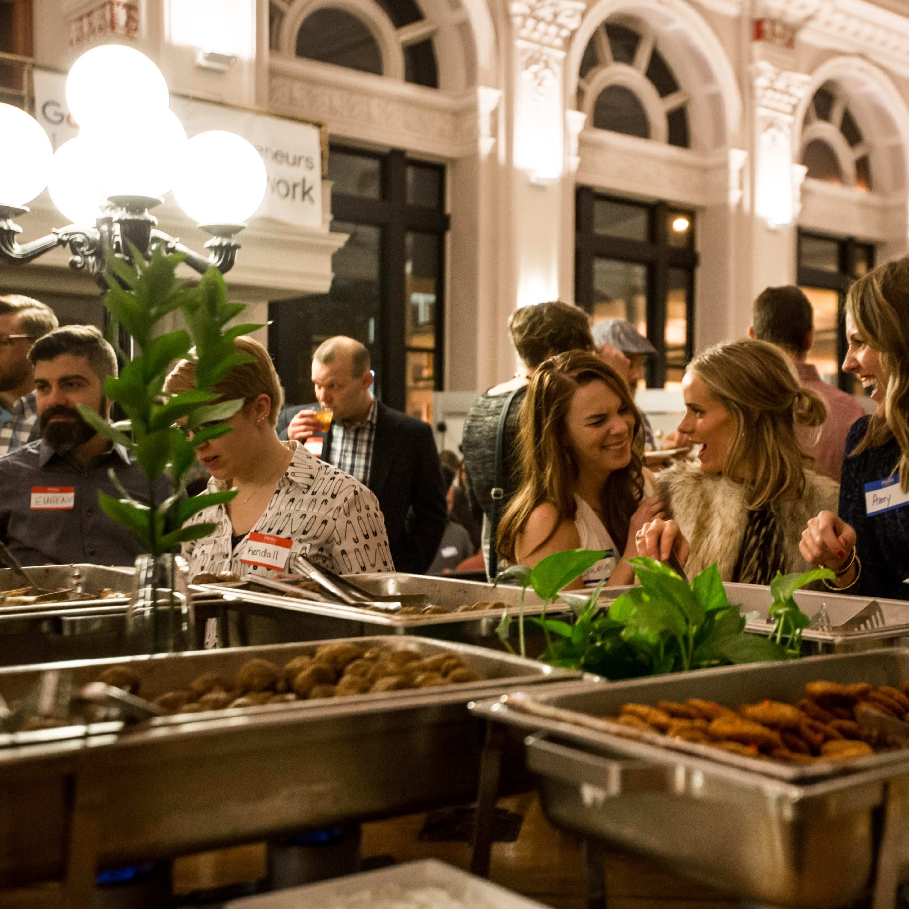 A group of people socializes at a buffet table filled with various food items in a well-lit indoor space.