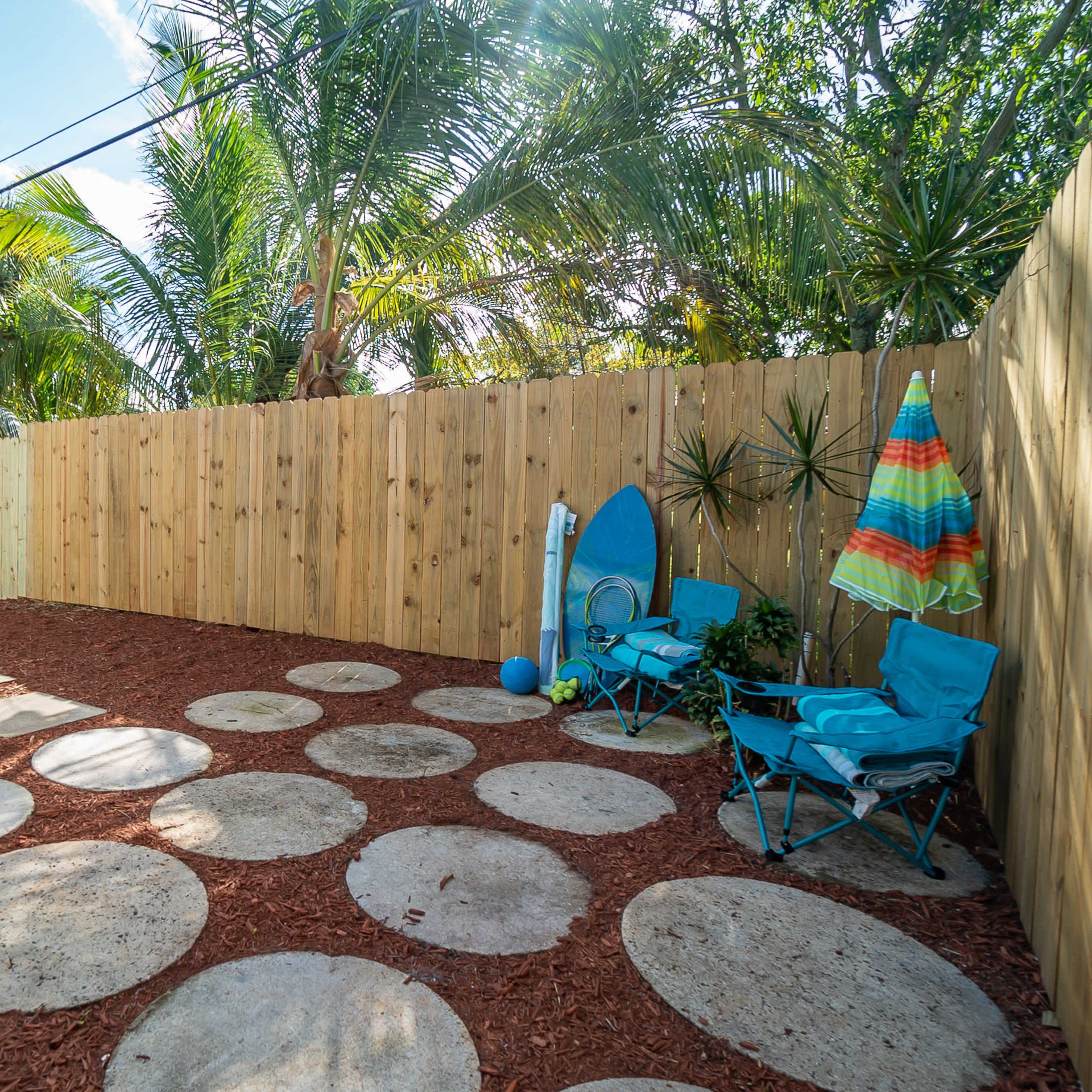 A landscaped backyard featuring a pathway of circular stone slabs surrounded by red mulch, with blue beach chairs and a surfboard leaning against a wooden fence.