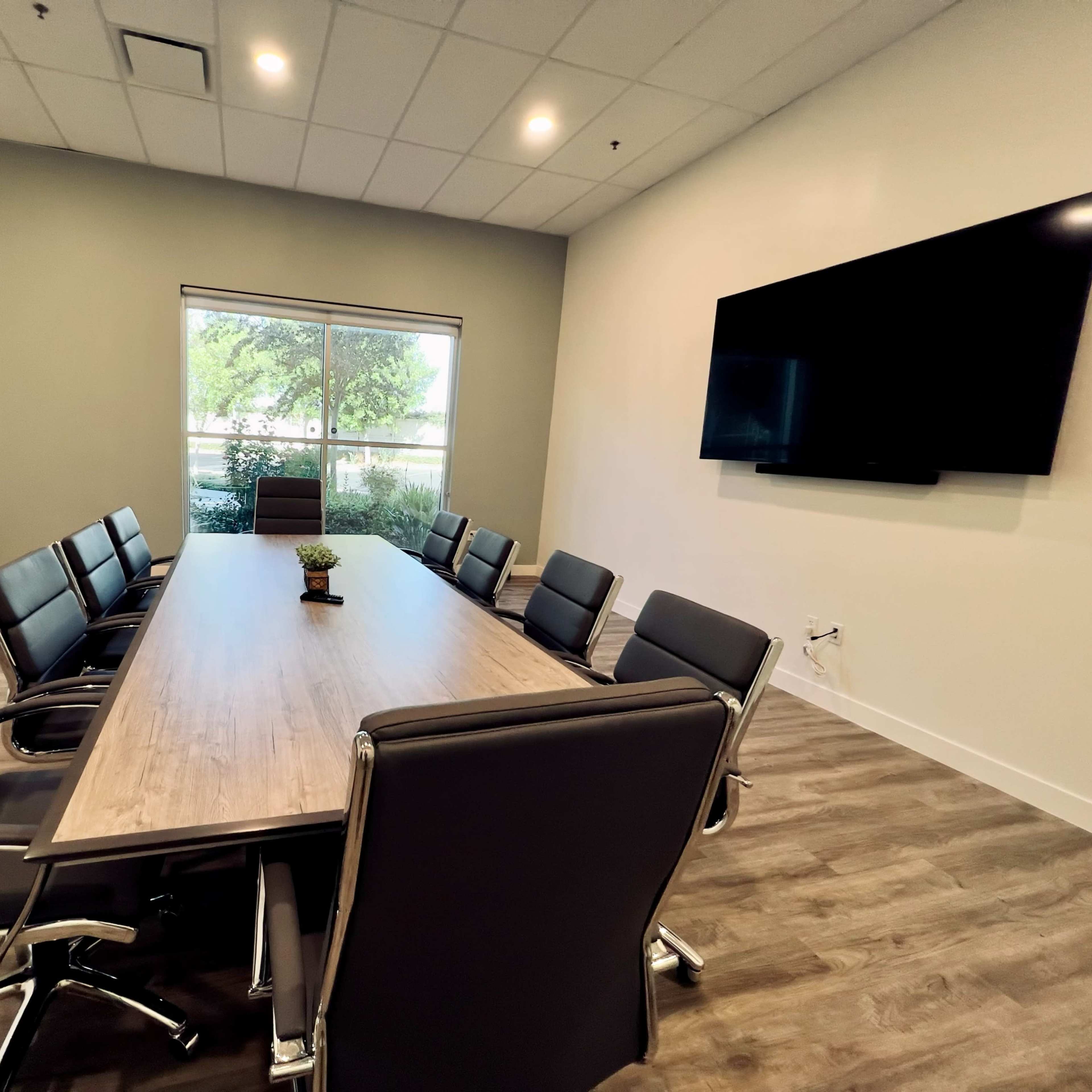 A conference room features a large wooden table surrounded by eight black chairs, with a television mounted on the wall and a window view of greenery outside.