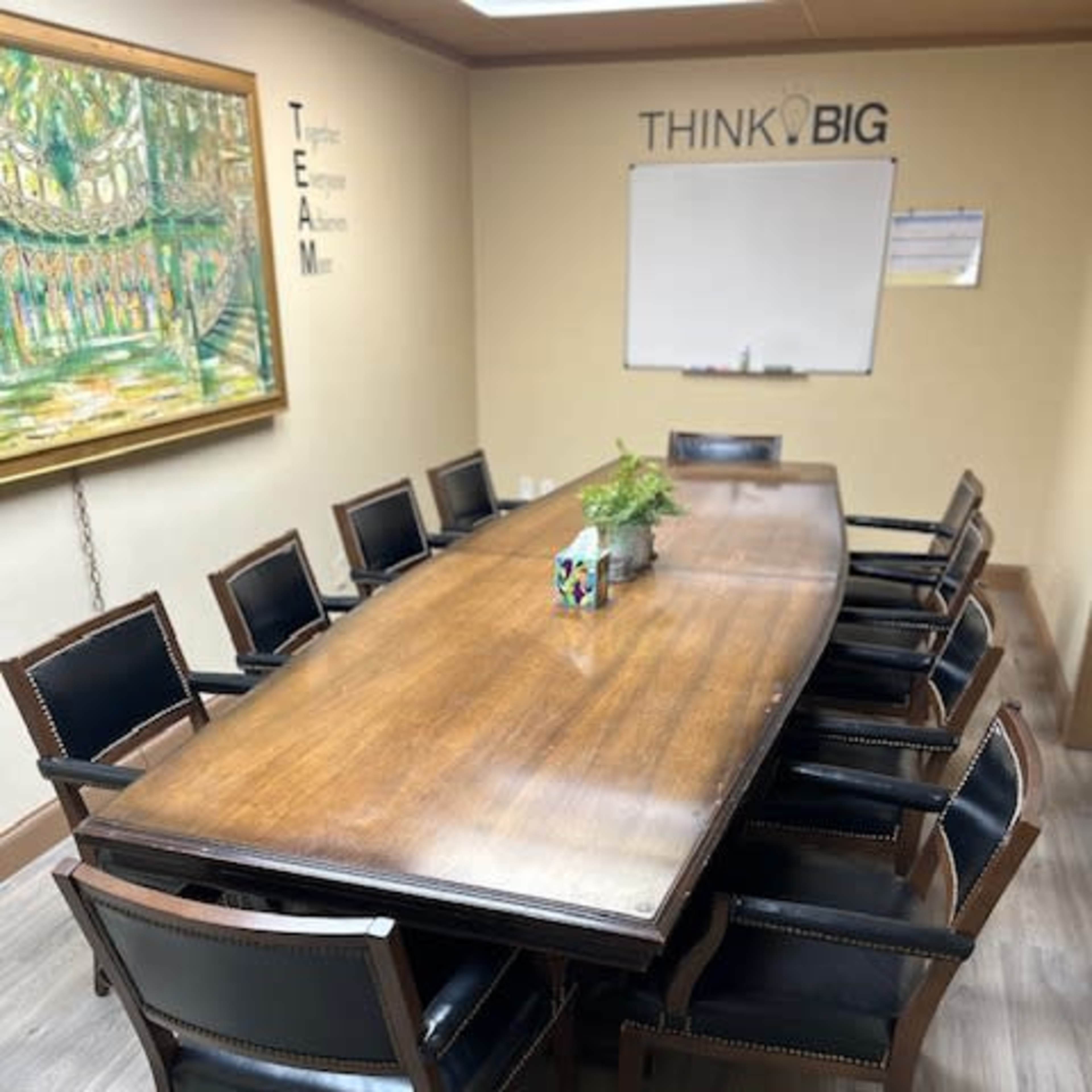 A long wooden conference table surrounded by black leather chairs is set in a meeting room with a whiteboard and a decorative painting on the wall.