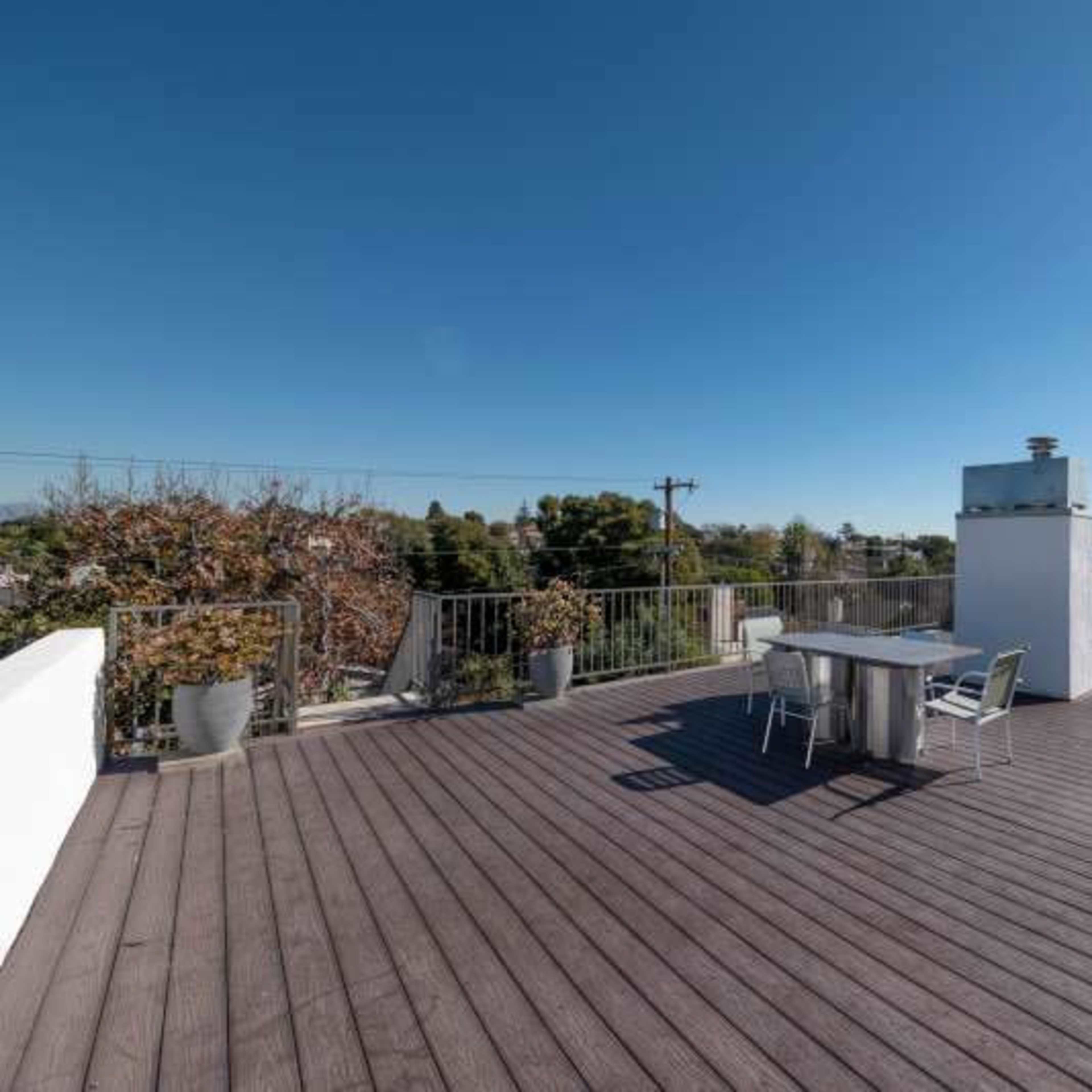 A spacious rooftop deck with wooden flooring, a barbecue grill, and a few chairs, surrounded by planters and a clear blue sky.