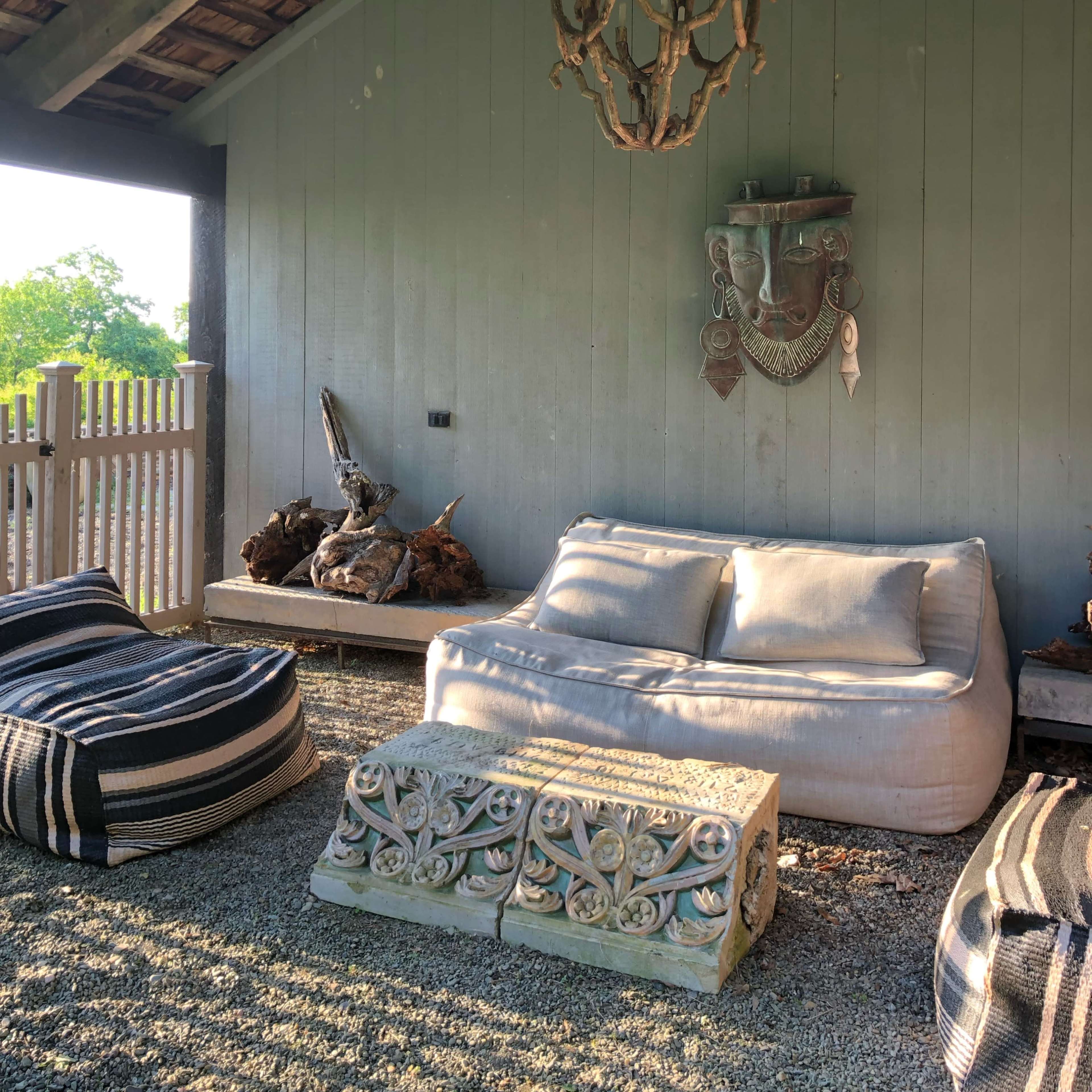 A cozy outdoor lounge area with a wooden ceiling, a decorative chandelier, and two large sofas on a gravel surface.