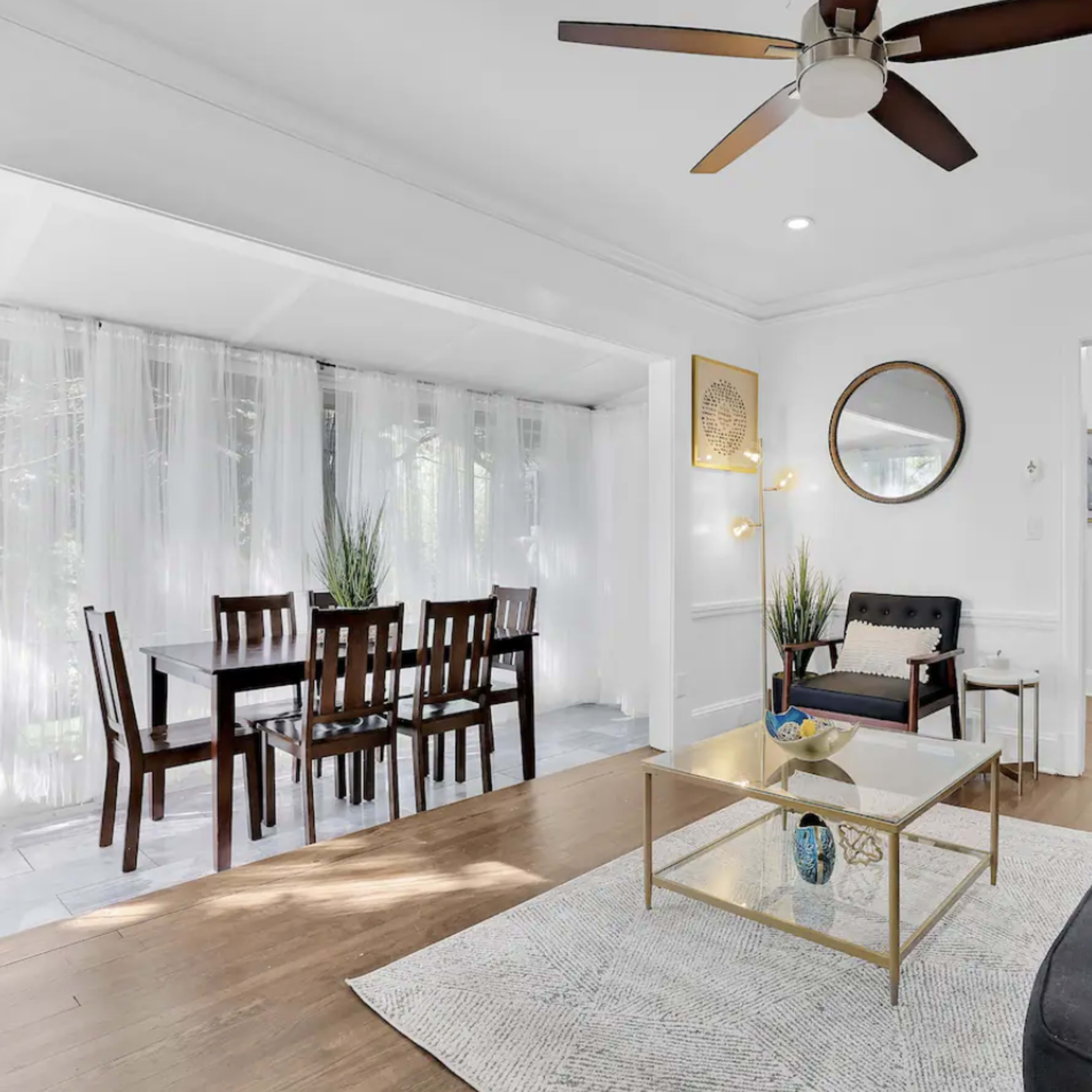 A bright, modern living room with a seating area, a dining table nearby, and large windows with sheer curtains allowing natural light to enter.