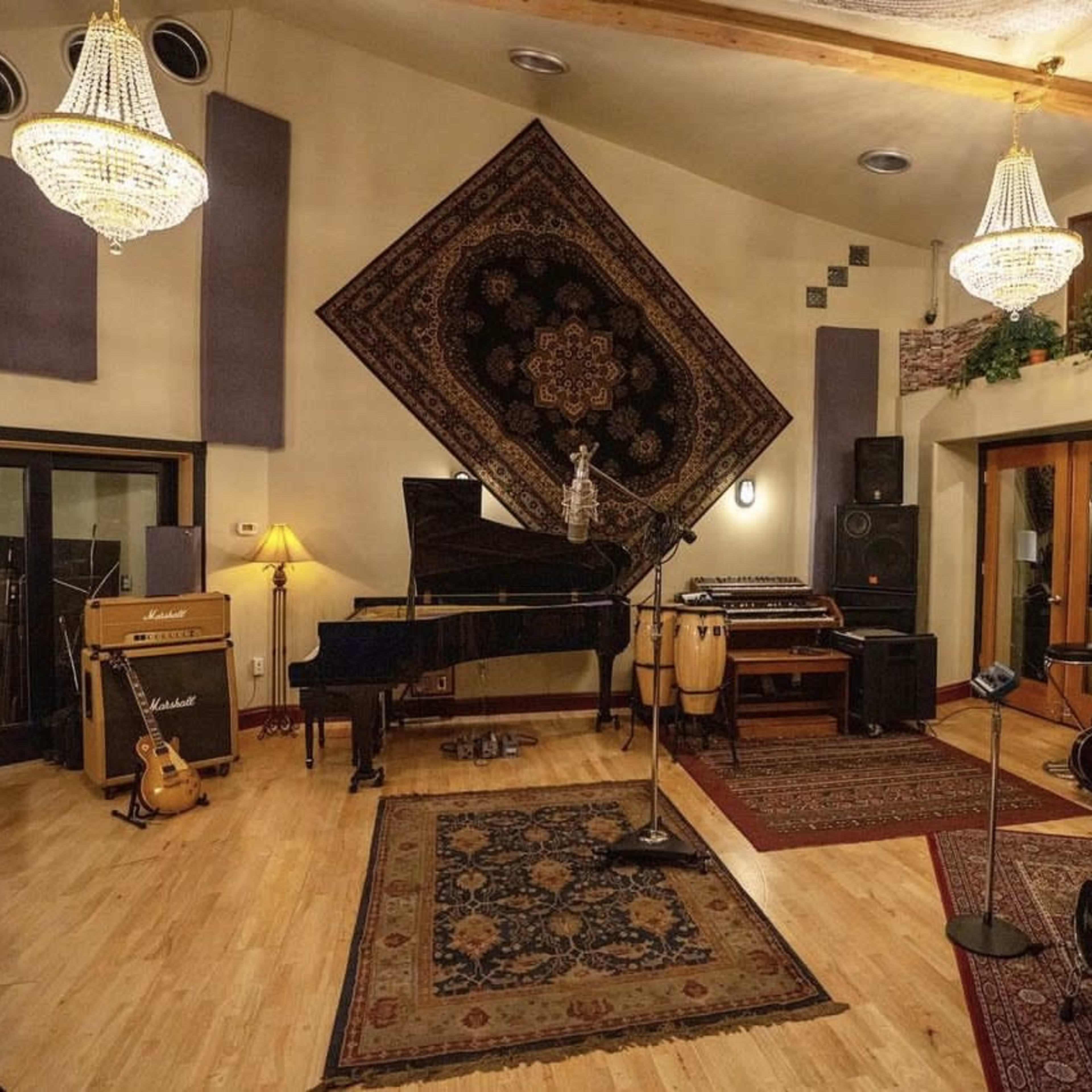 A music studio featuring a grand piano, various instruments, sound equipment, and decorative rugs under decorative lighting fixtures.