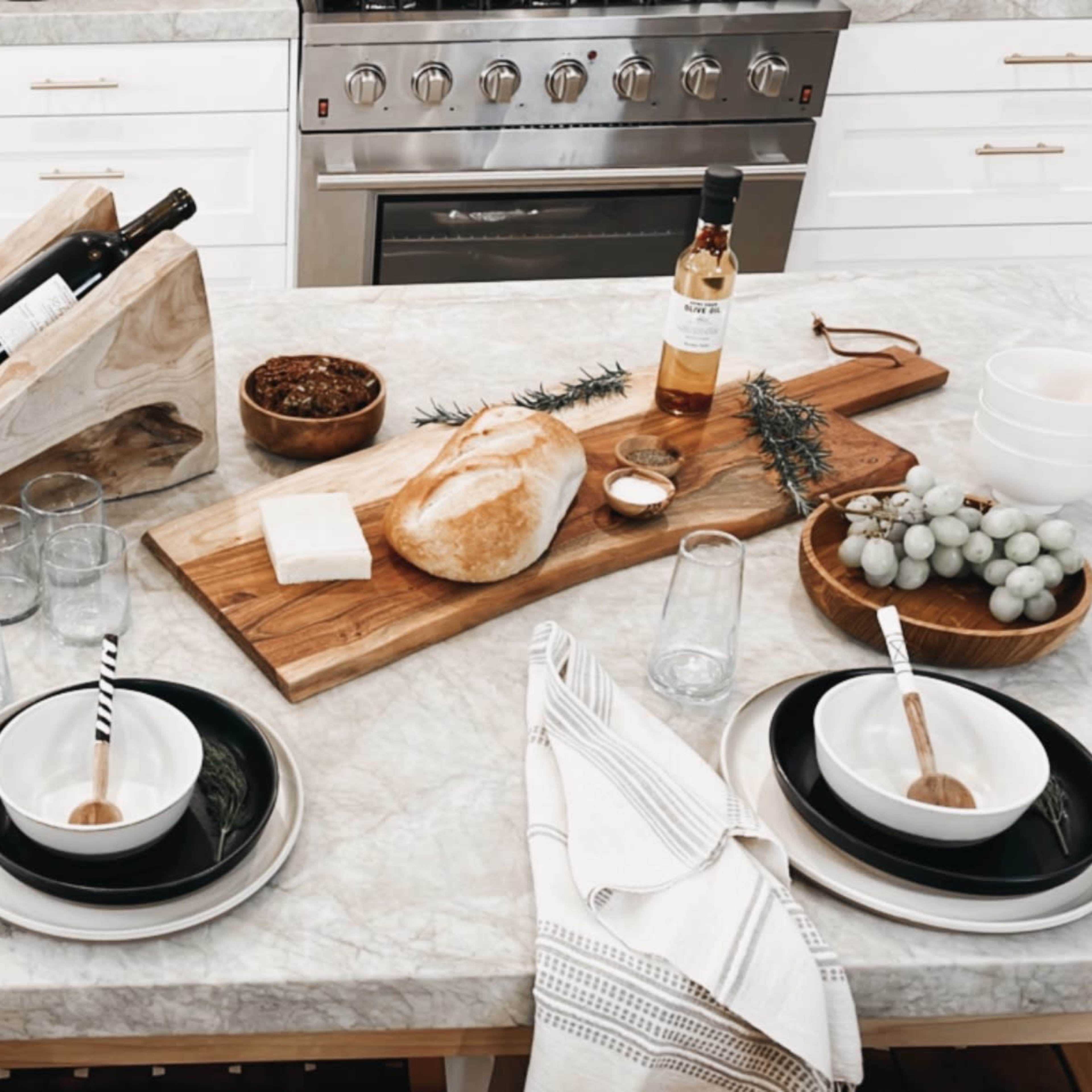 A kitchen table is set with black and white dishware, glasses, and a variety of food items, including bread, raspberries, and a bottle of wine, arranged on a marble countertop.