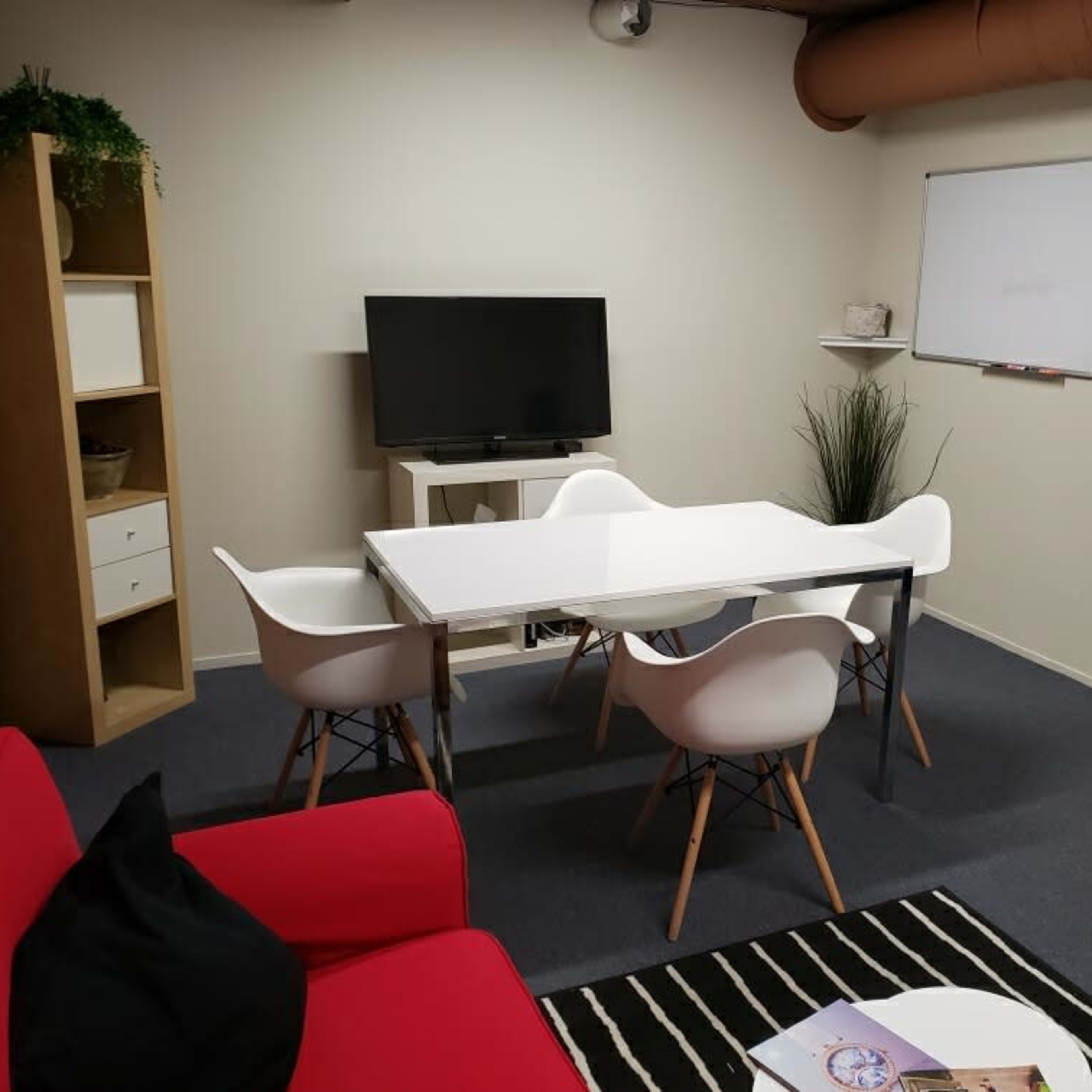 A small meeting room with a white table surrounded by four chairs, a television, a whiteboard, and a shelf plant.