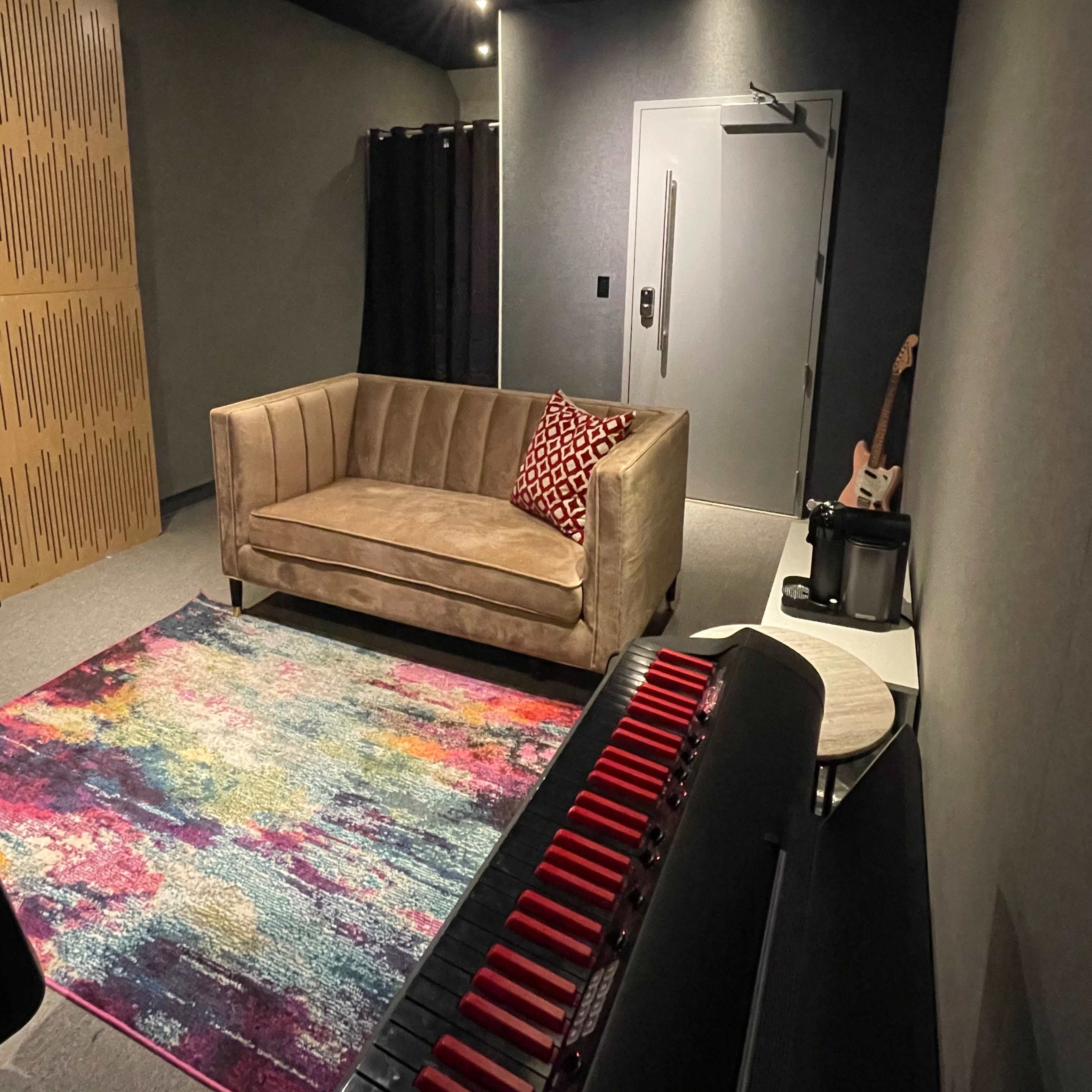 A cozy music studio features a sofa, colorful area rug, and a keyboard in a dimly lit room with soundproof walls.