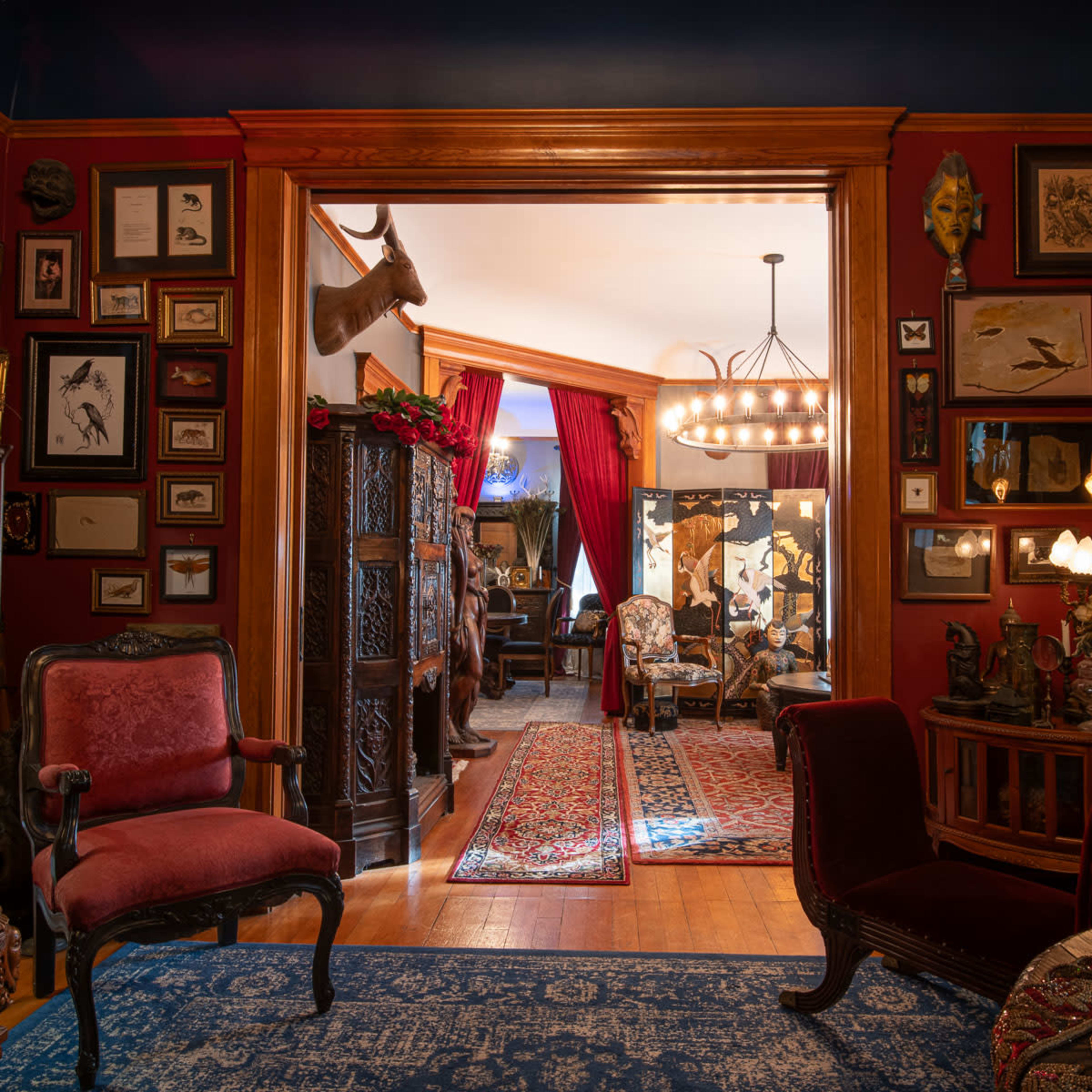 A richly decorated room features ornate wooden furniture, framed wall art, and a view into another elegantly furnished space.