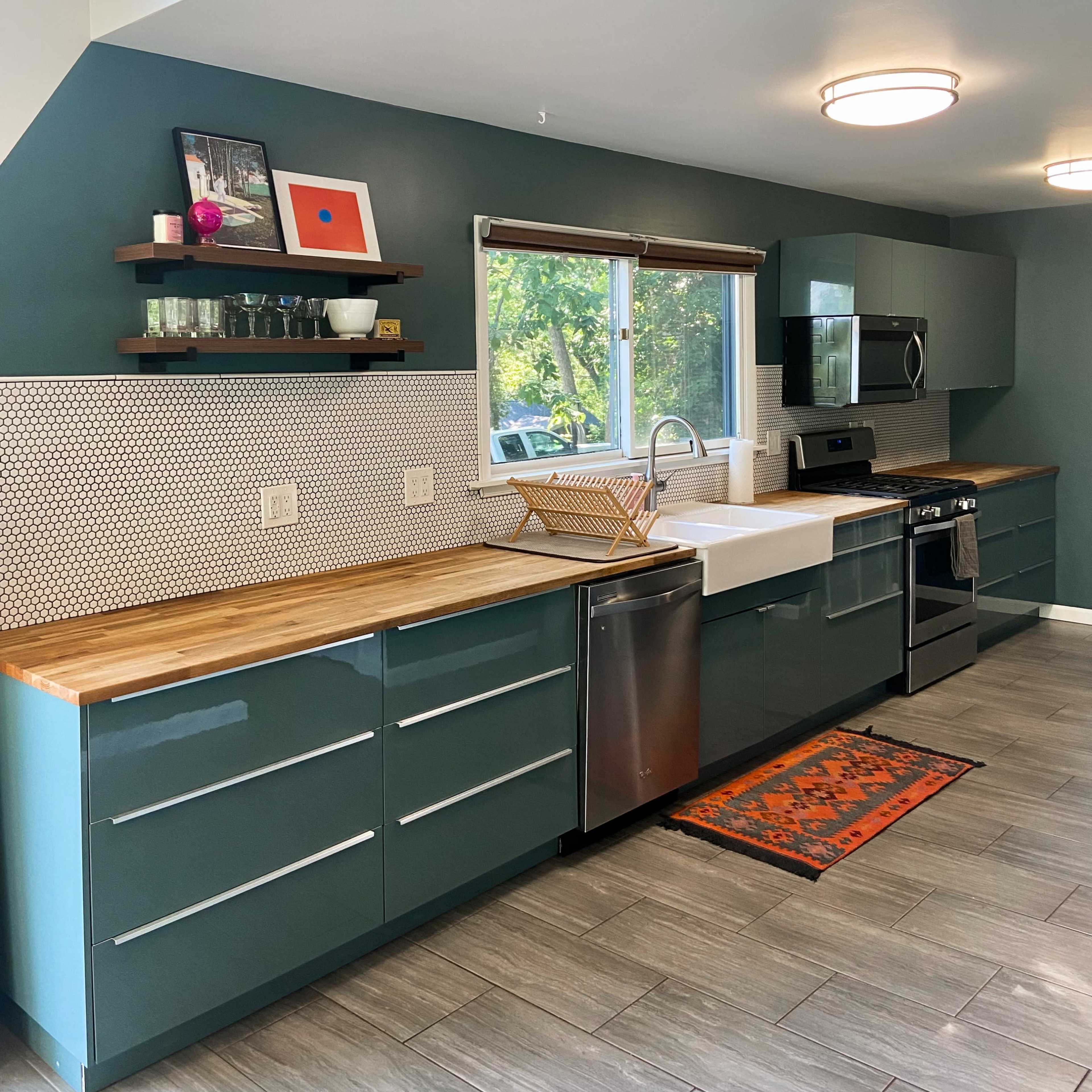 A modern kitchen with teal cabinets, a stainless steel refrigerator, a white sink, and wood accents, featuring a window with natural light.