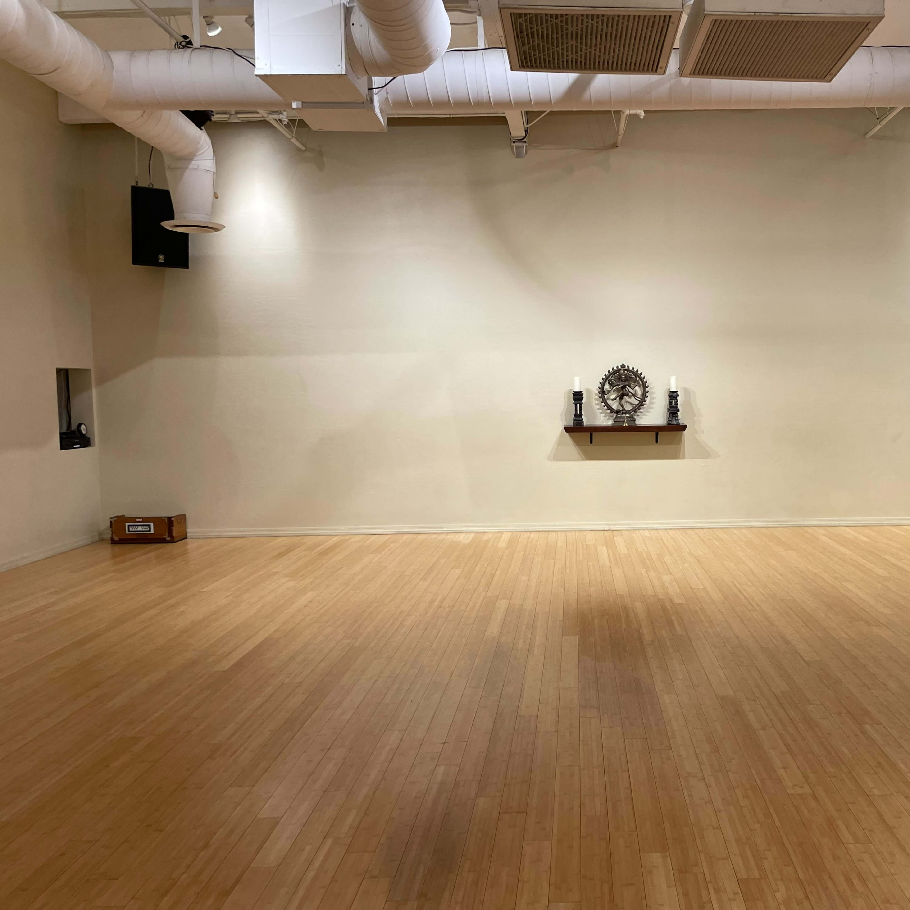 An empty room with wooden flooring, featuring a wall-mounted shelf displaying a clock, and two speakers mounted on the walls.
