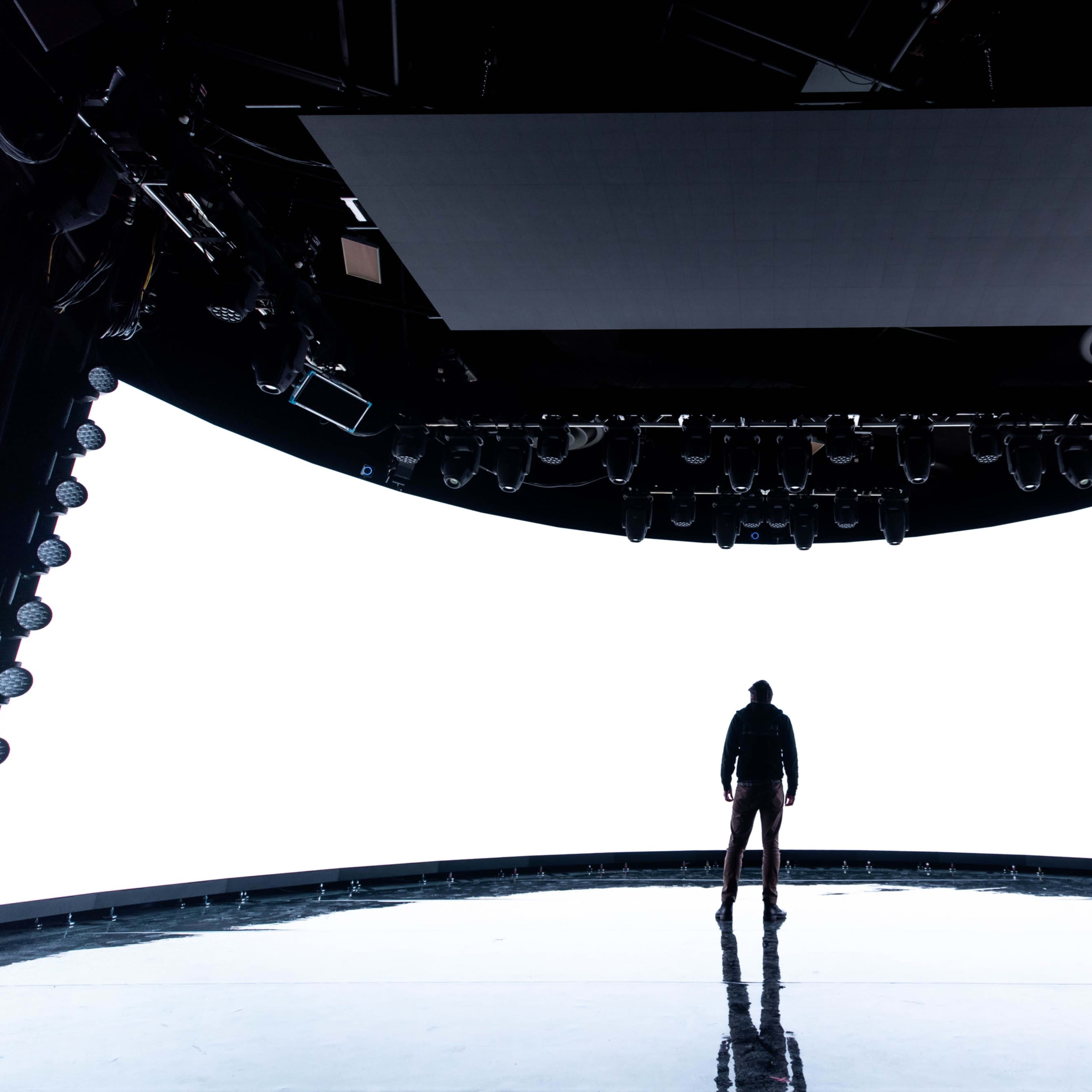 A person stands alone on a dimly lit stage with a large blank screen behind them.