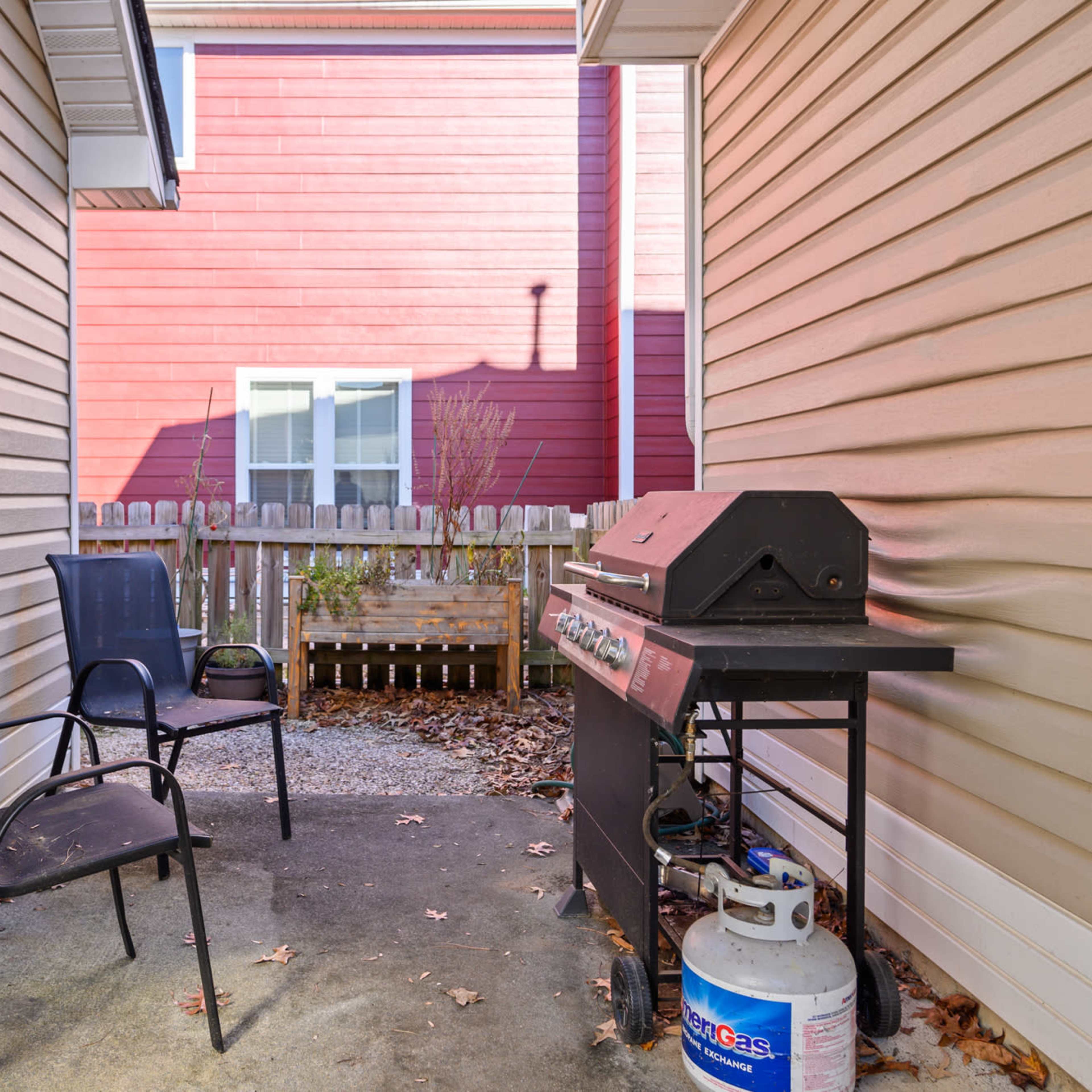 A small outdoor area between two houses, featuring a grill and two black chairs on a concrete surface, with a wooden fence and a glimpse of greenery in the background.