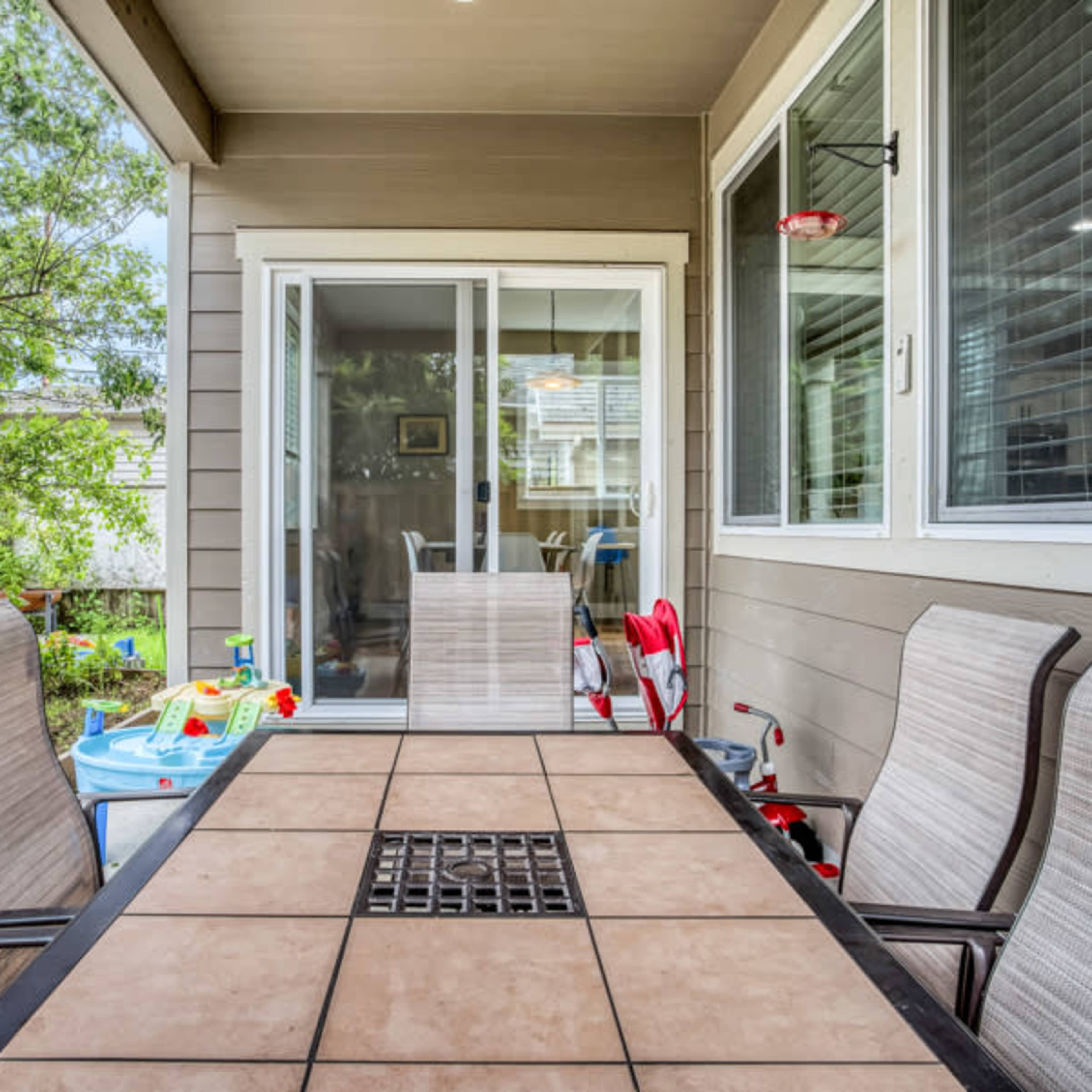 A patio area with a tiled table surrounded by four chairs, leading to a shaded porch with sliding glass doors and a view of a grassy yard.