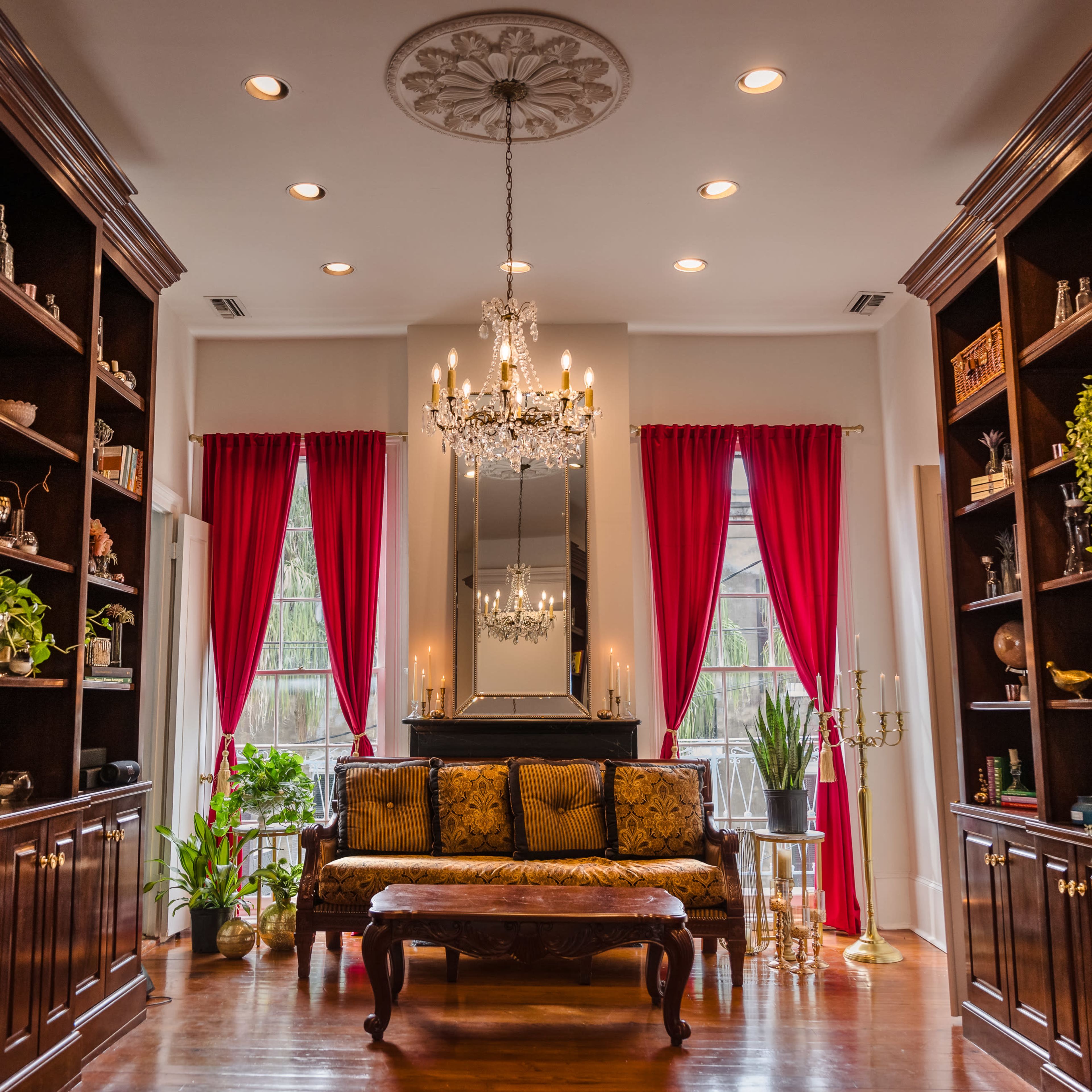 An elegantly decorated living room with dark wooden shelves, a central chandelier, and a plush sofa surrounded by plants and framed photos.