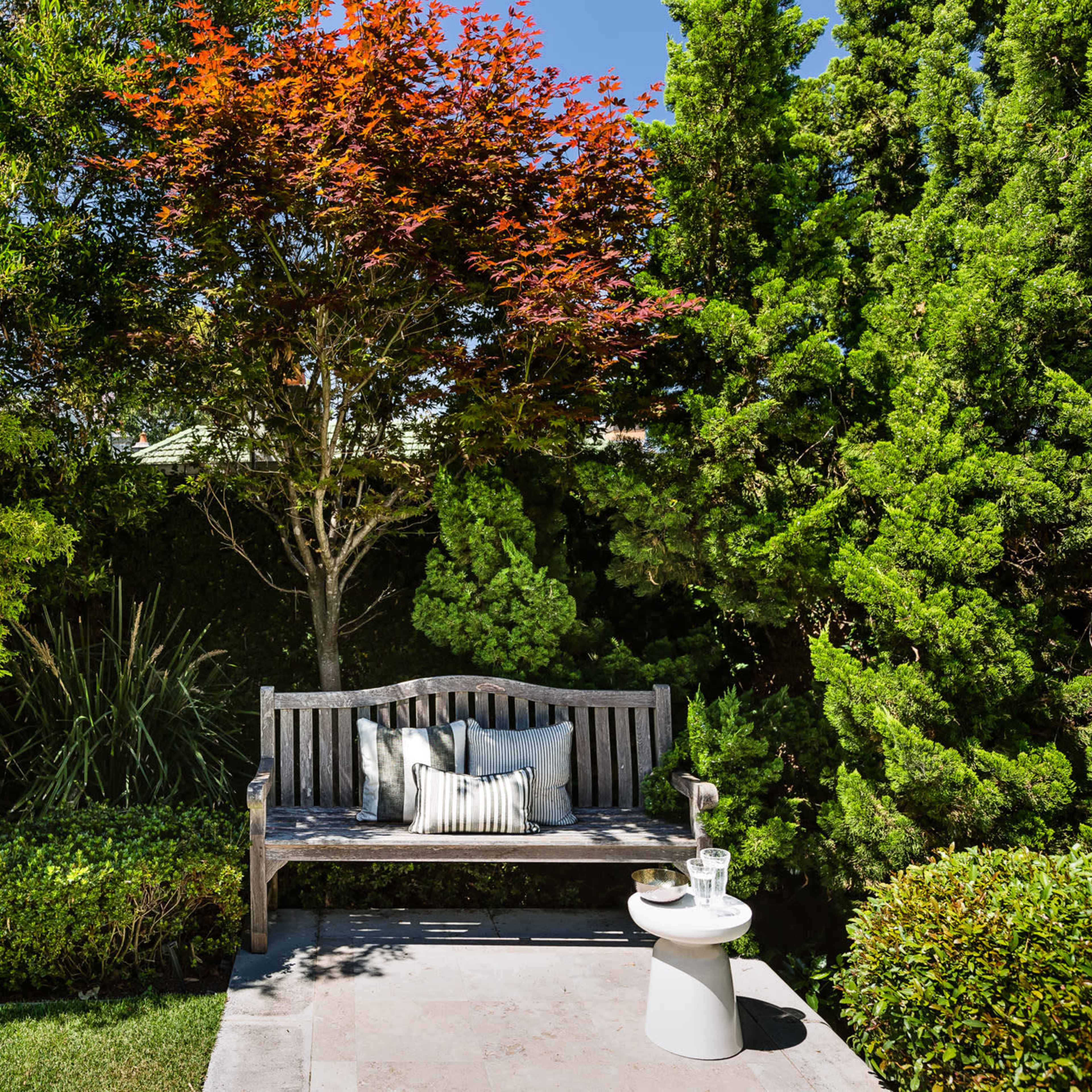 A wooden bench with a decorative pillow sits beside a small side table in a landscaped garden with vibrant trees and shrubs.
