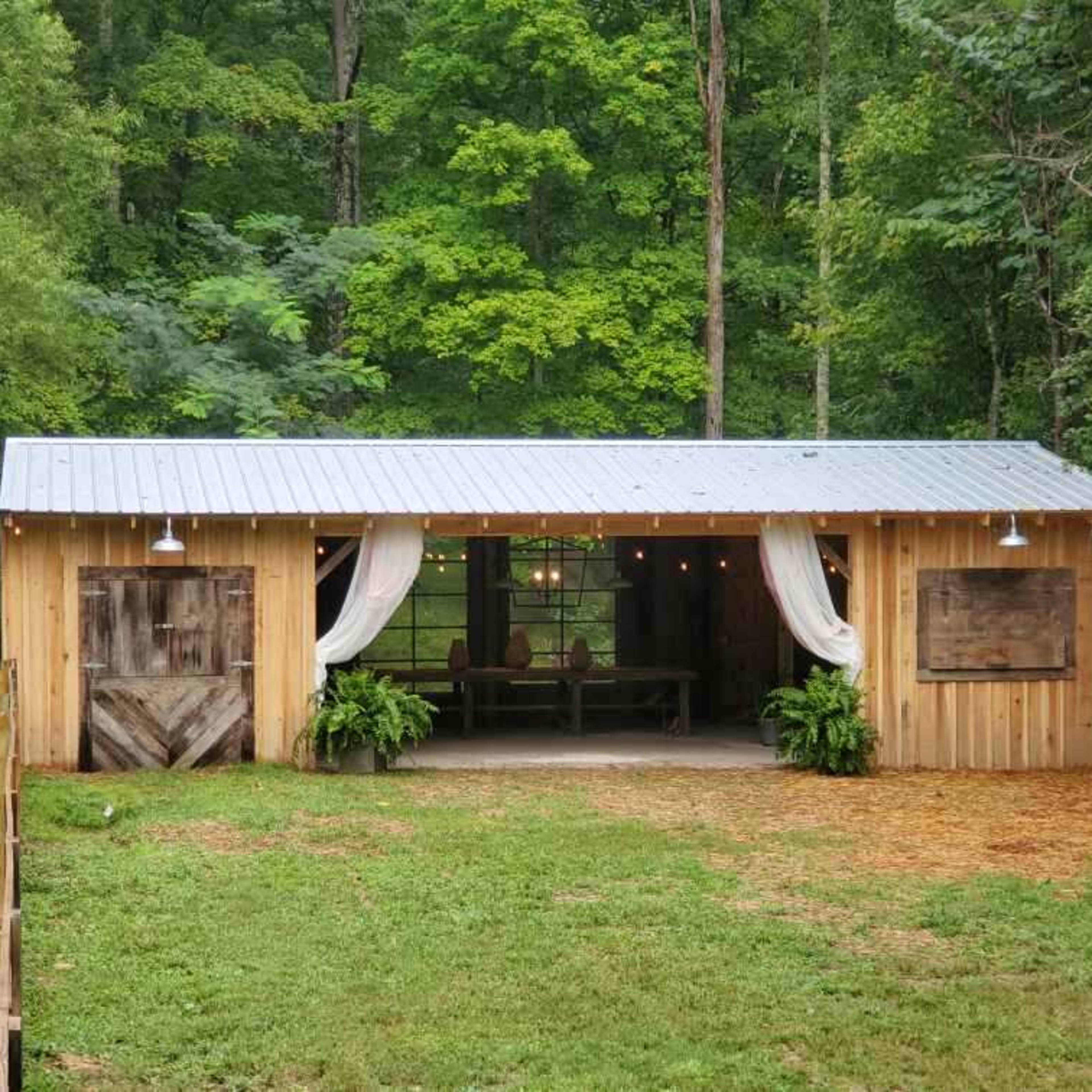 A wooden cabin with a metal roof features open sides, decorative curtains, and is surrounded by greenery.