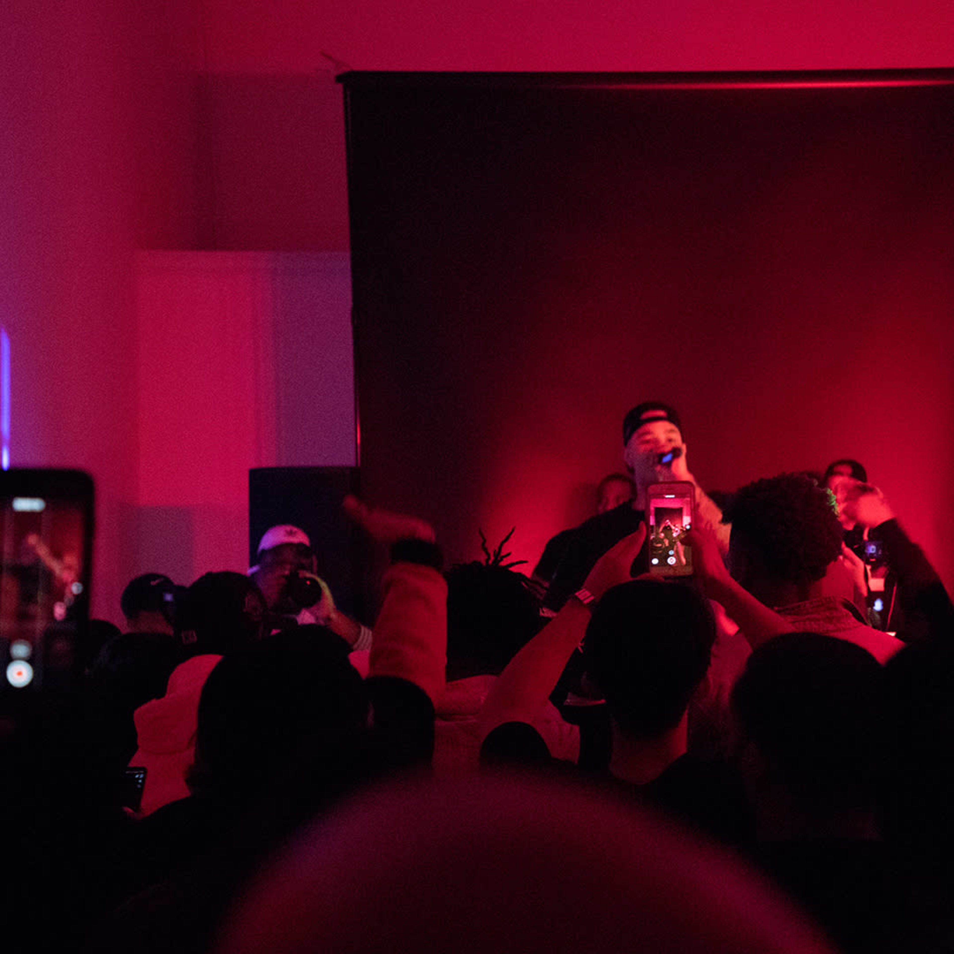 A crowd of people is gathered in a dimly lit venue with red lighting, watching a performer on stage while capturing the moment with their phones.