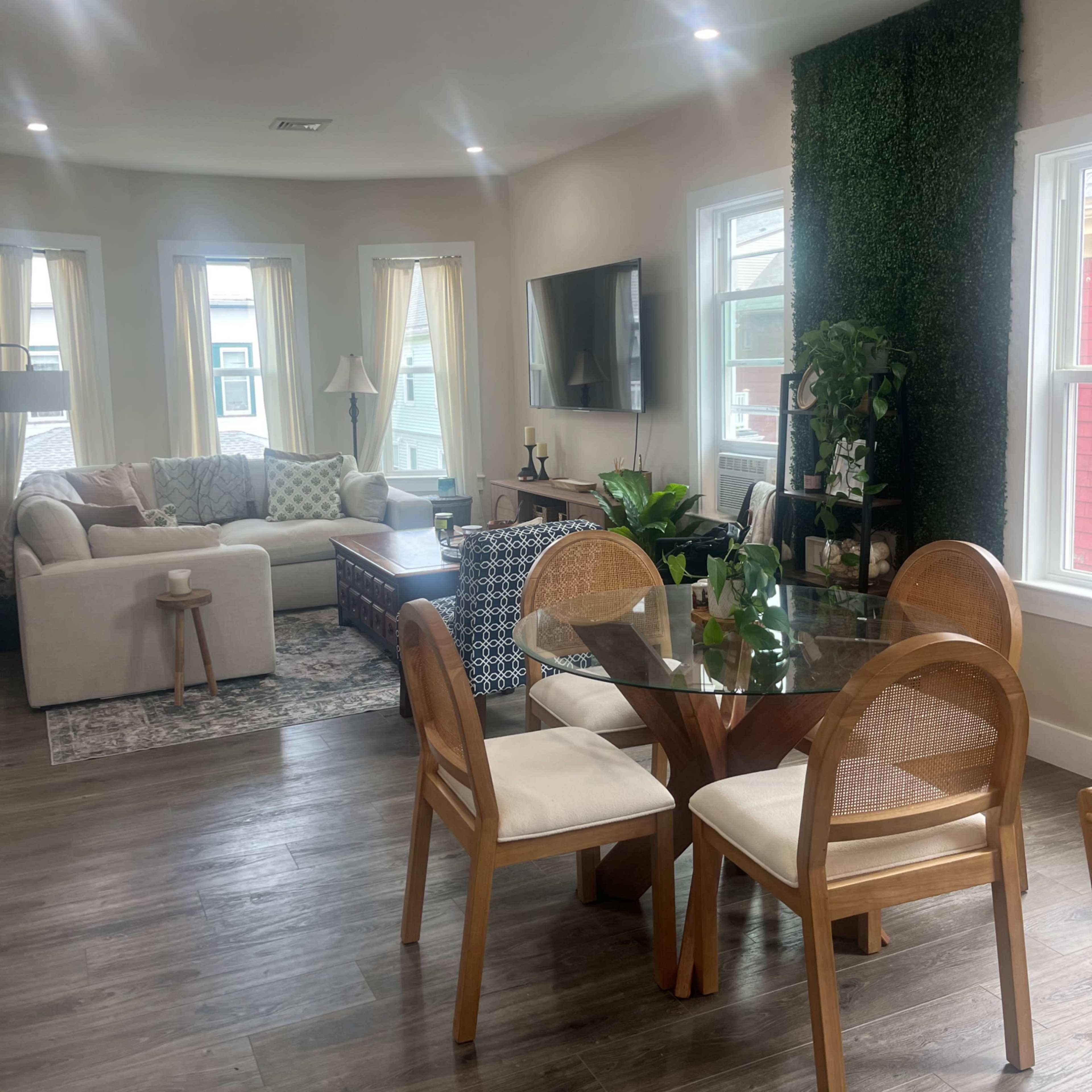 A bright living room features a cozy seating area with a sectional sofa and a glass dining table surrounded by wooden chairs.