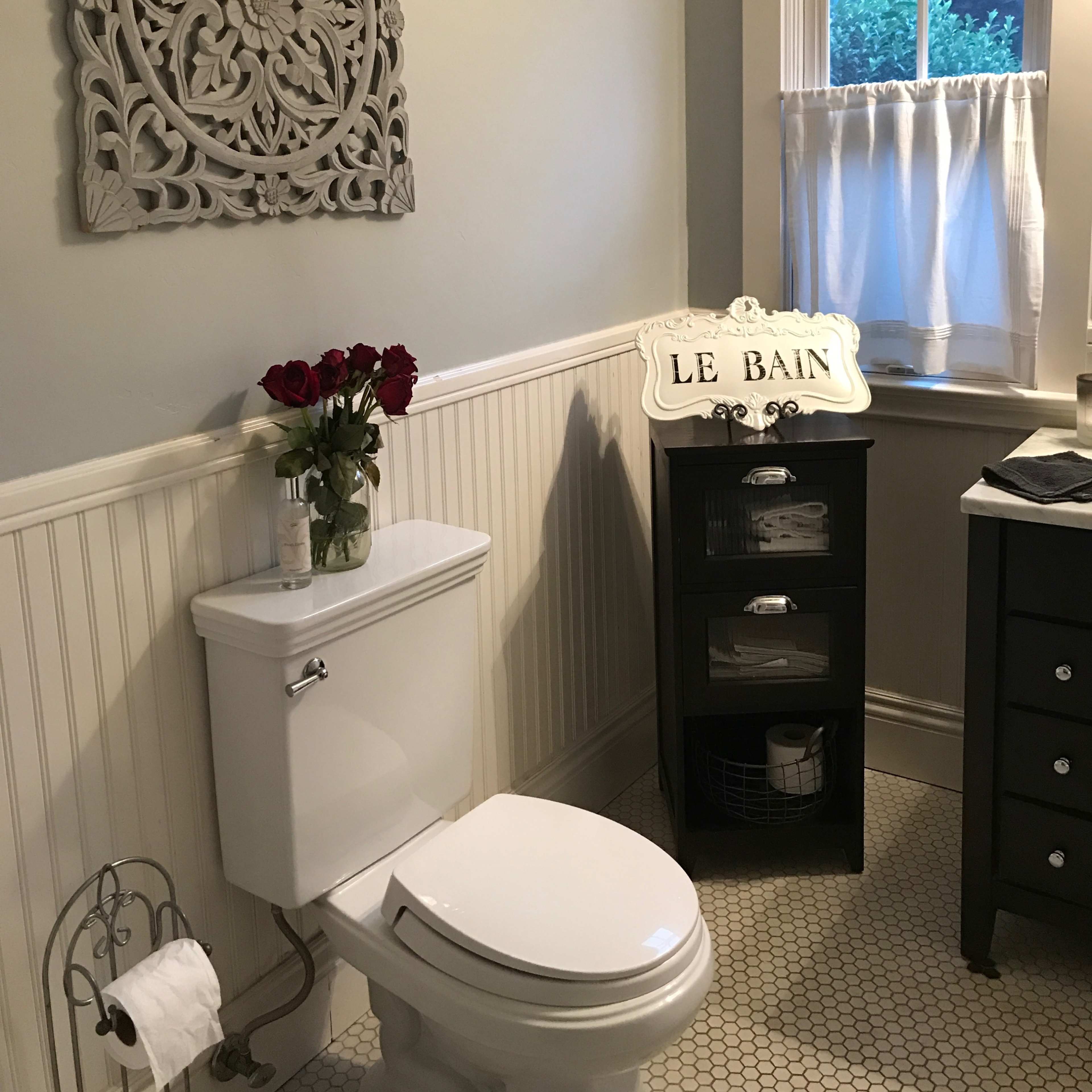 A bathroom with a white toilet, a decorative wall piece, a window with white curtains, a vase of roses, and a black storage cabinet.
