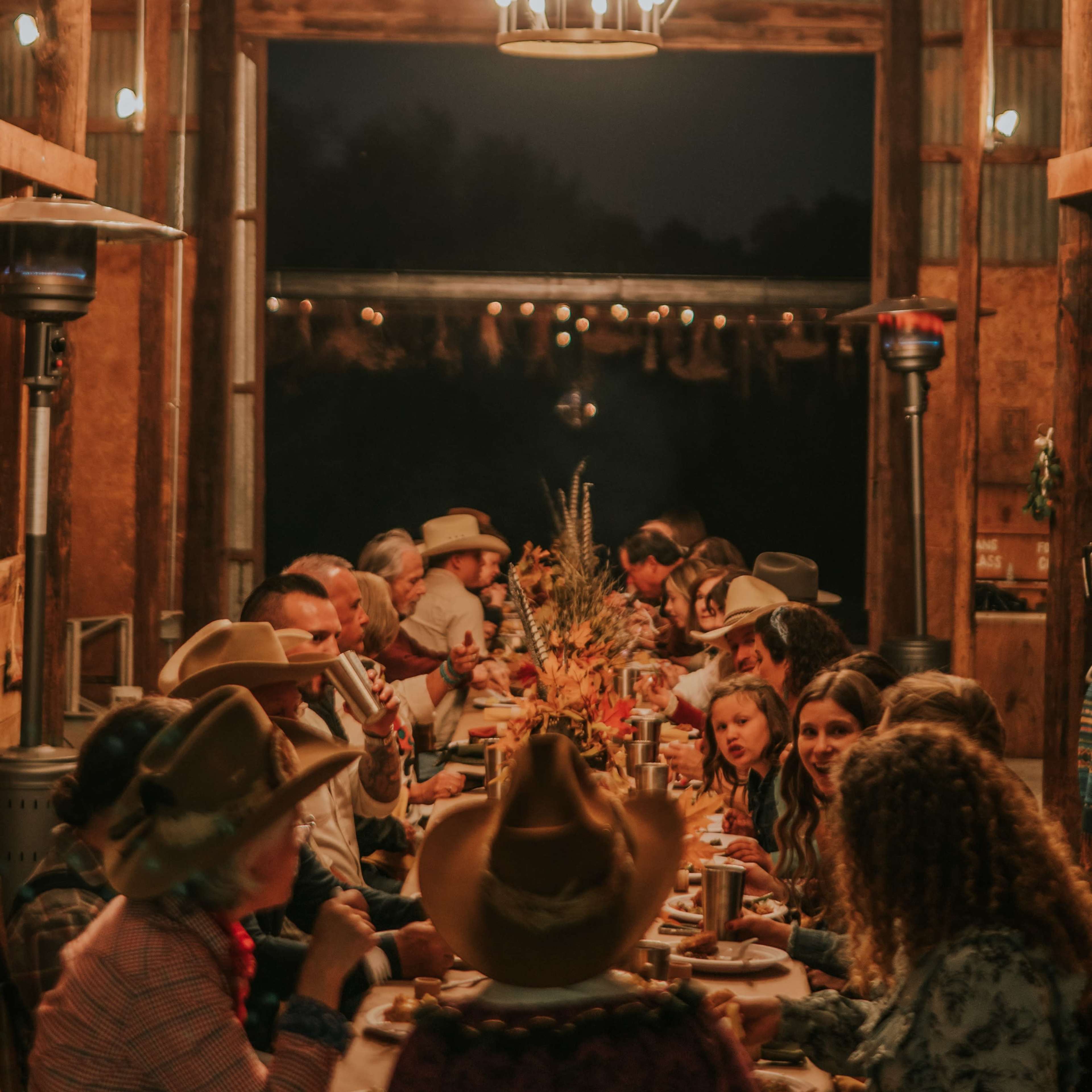 A long table set for a dinner gathering is illuminated by large circular chandeliers in a rustic barn at night.