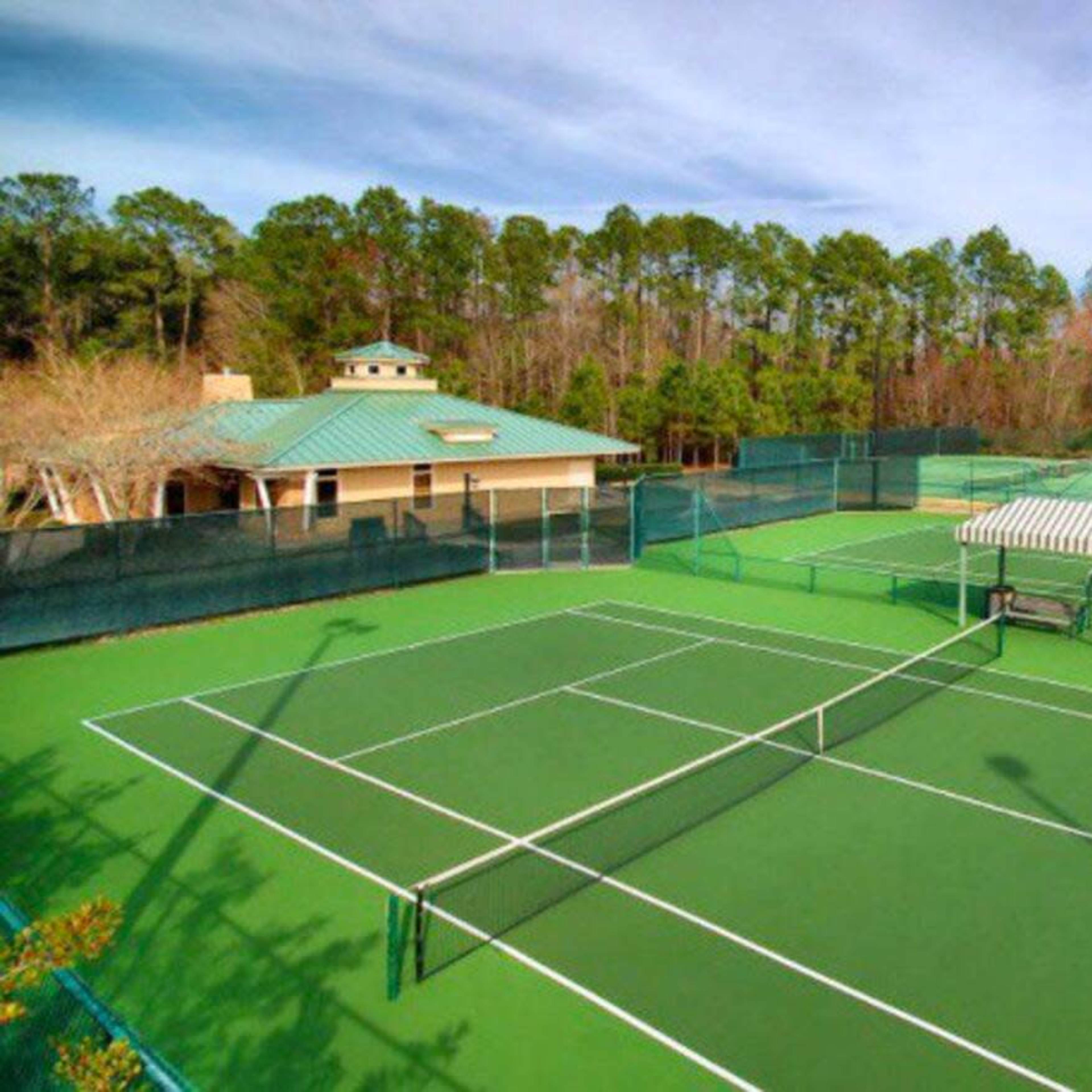 A tennis facility with multiple green courts surrounded by trees and two buildings in the background.