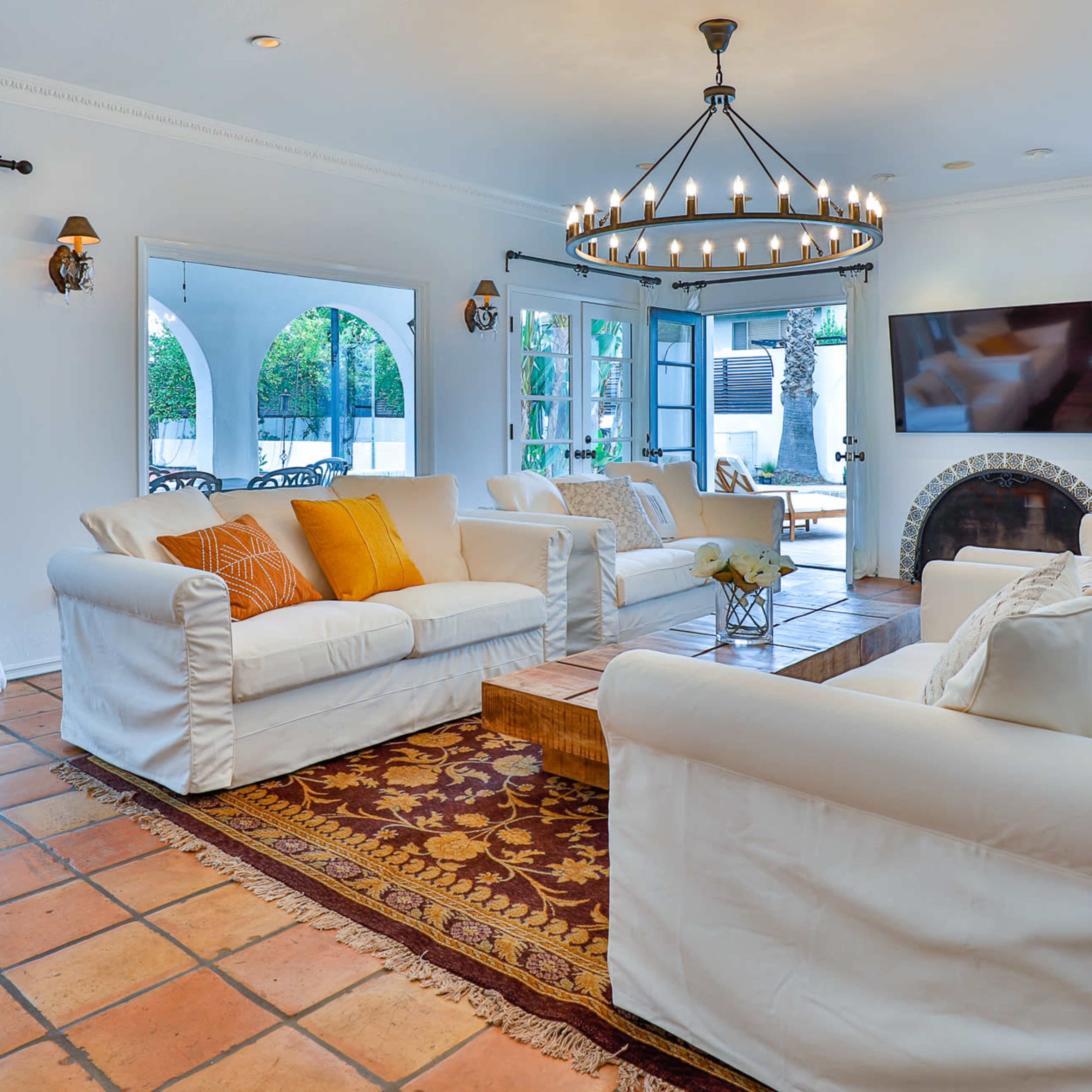 A bright living room with white sofas, a wooden coffee table, and a large chandelier, framed by arched doorways leading to a patio.