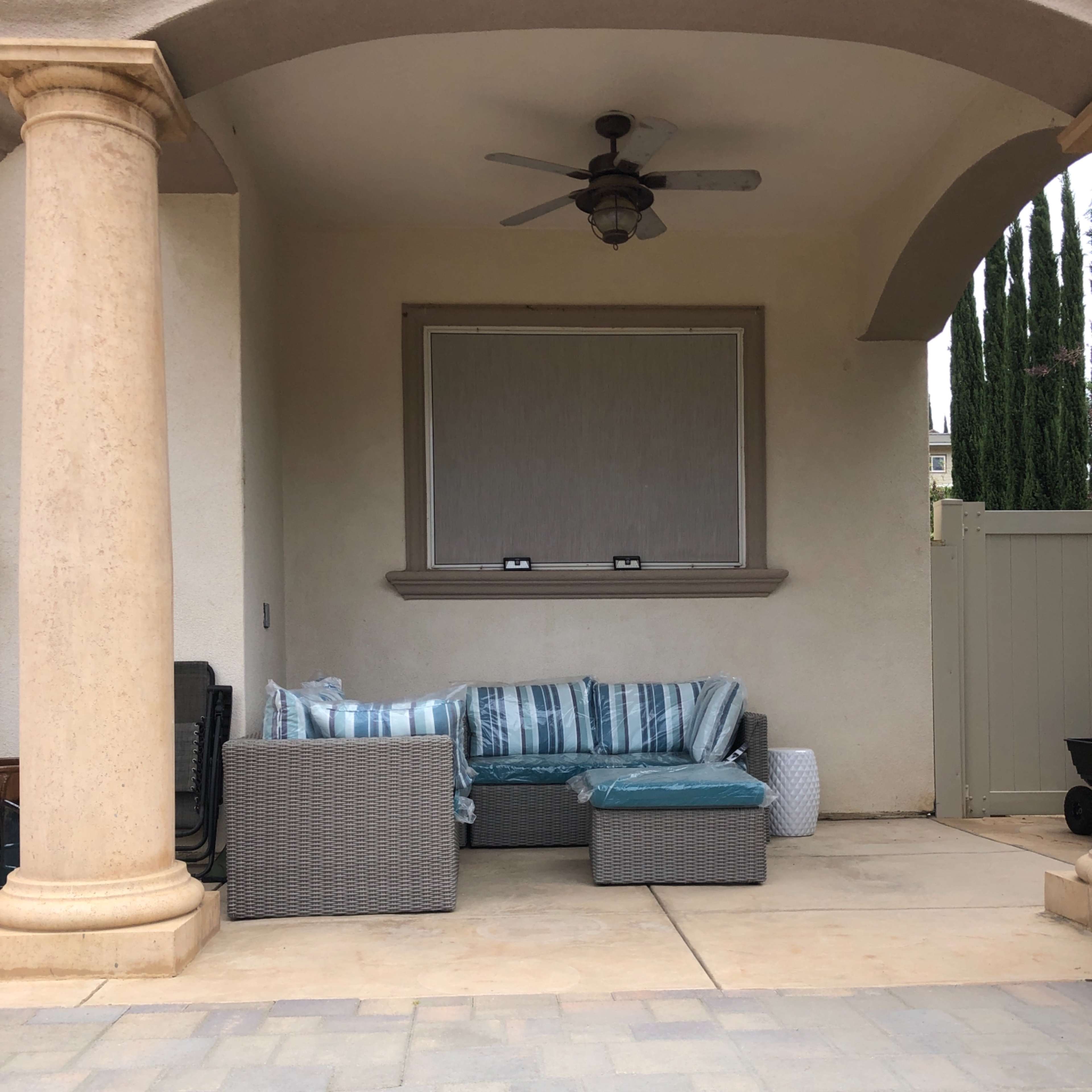A covered outdoor seating area featuring a sectional couch with blue and white striped cushions, flanked by stone columns and a fan overhead.