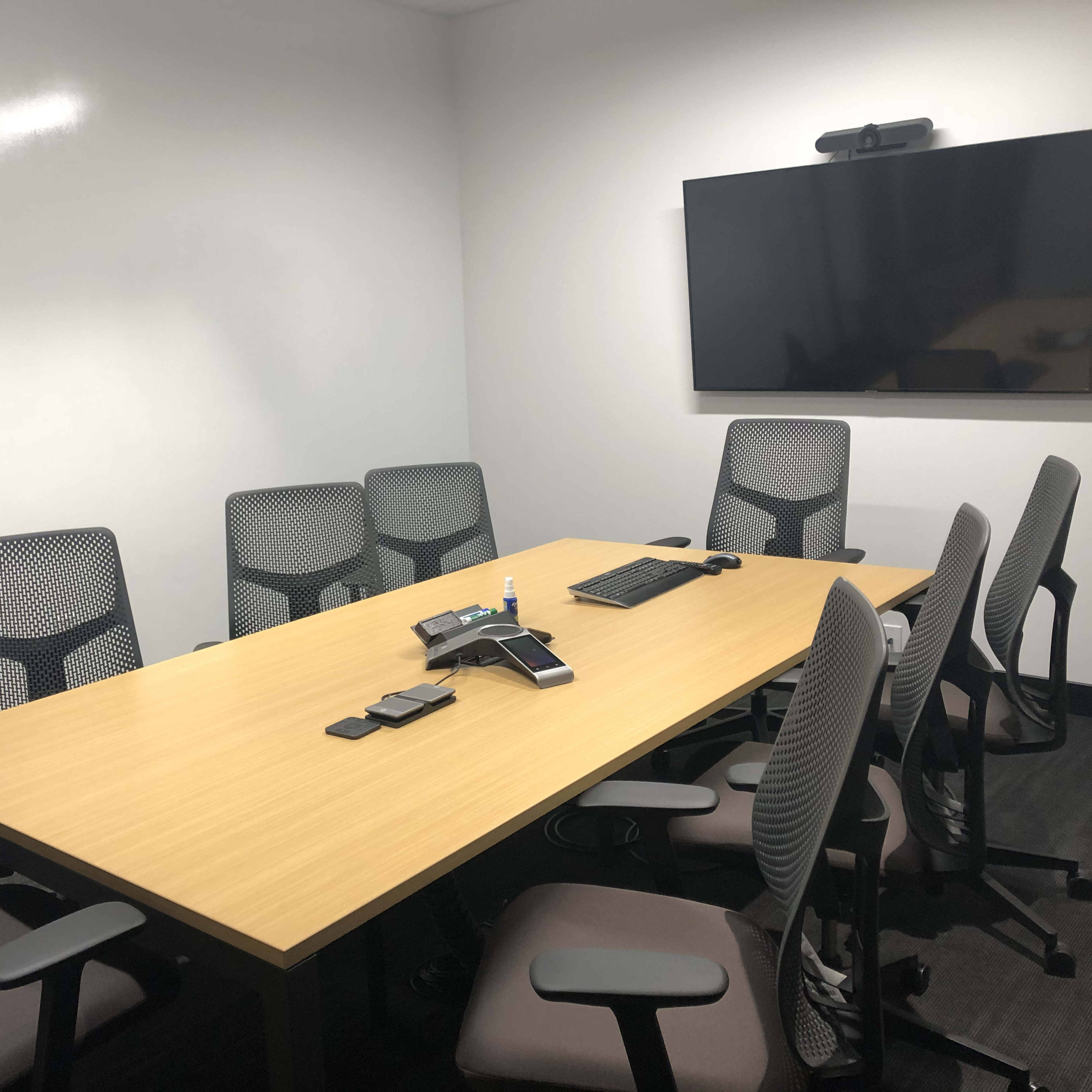 A conference room is set up with a large wooden table surrounded by eight mesh office chairs, equipped with a phone and a monitor on the wall.