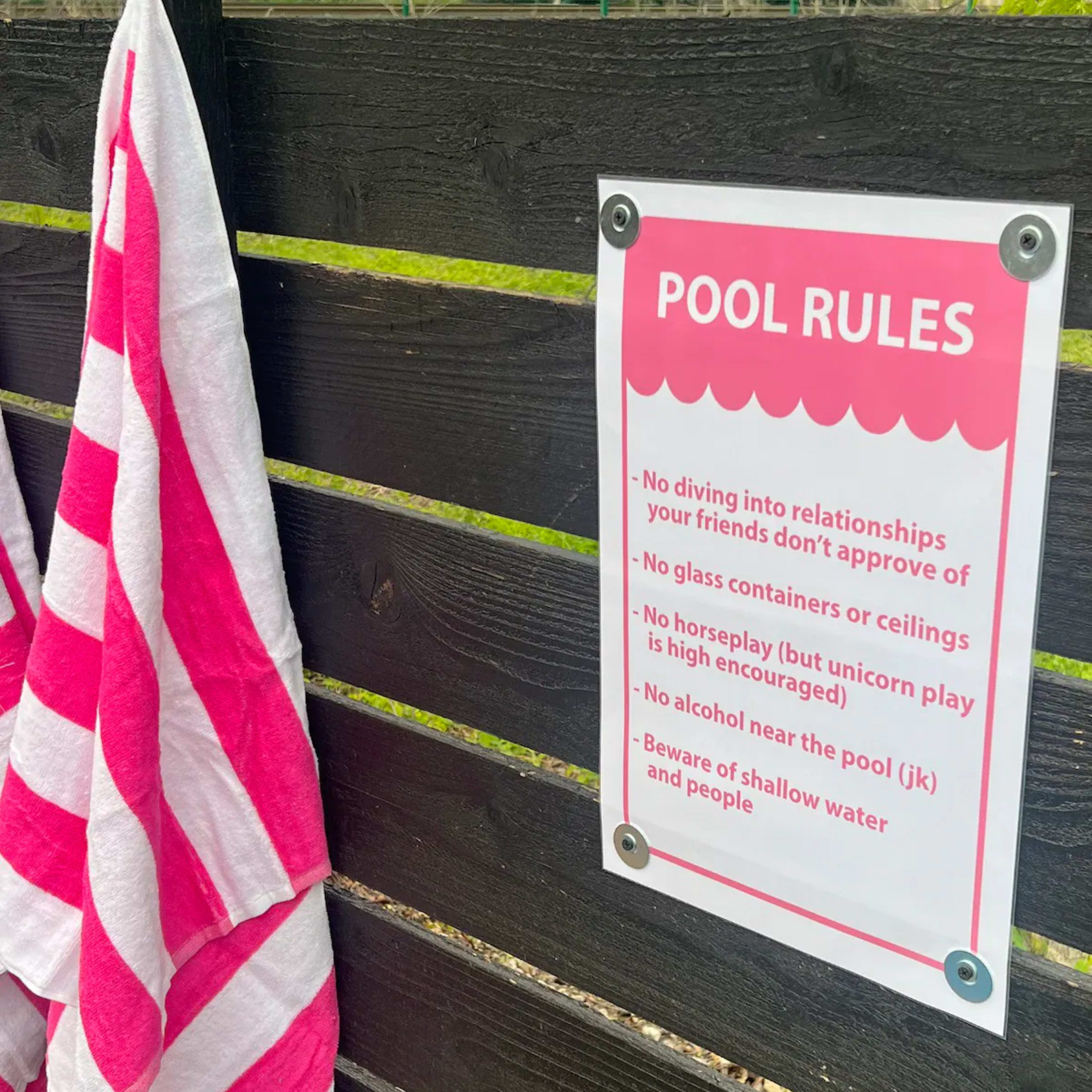 A sign detailing humorous pool rules hangs on a black wooden fence next to a pink-striped towel.