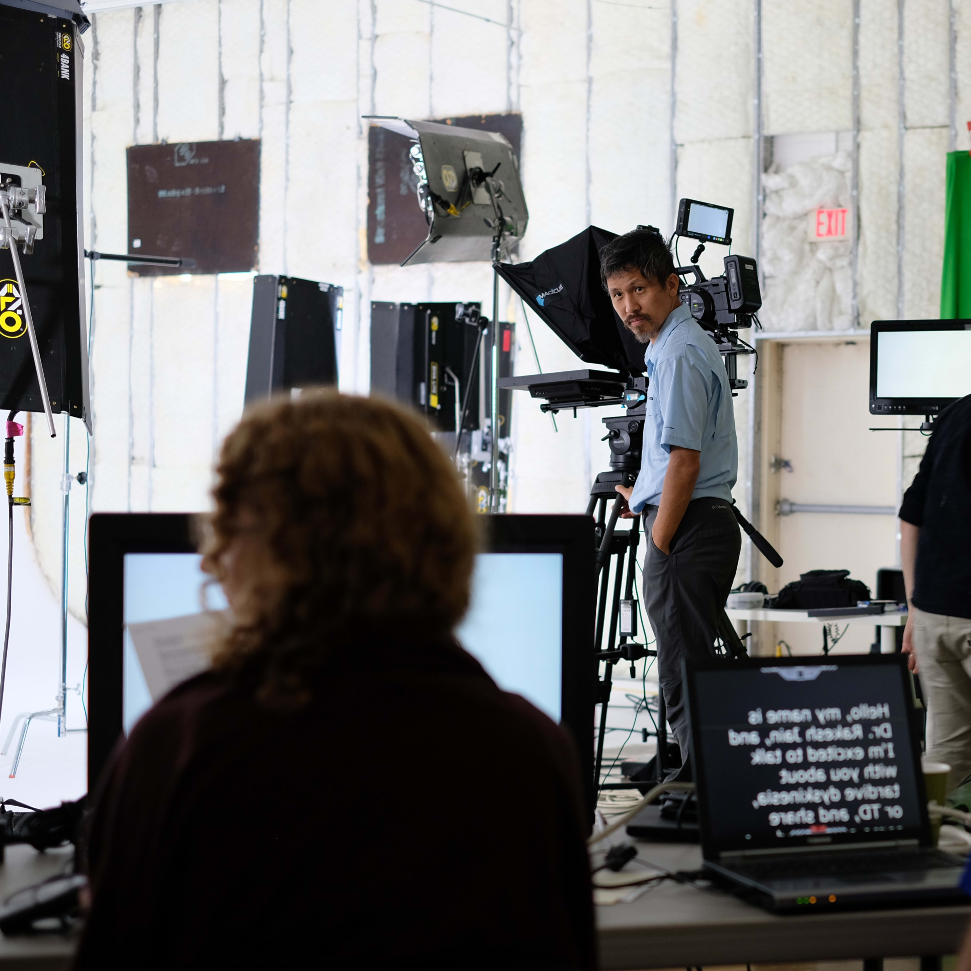 A studio setup features cameras and monitors, with crew members operating equipment and preparing for a shoot.