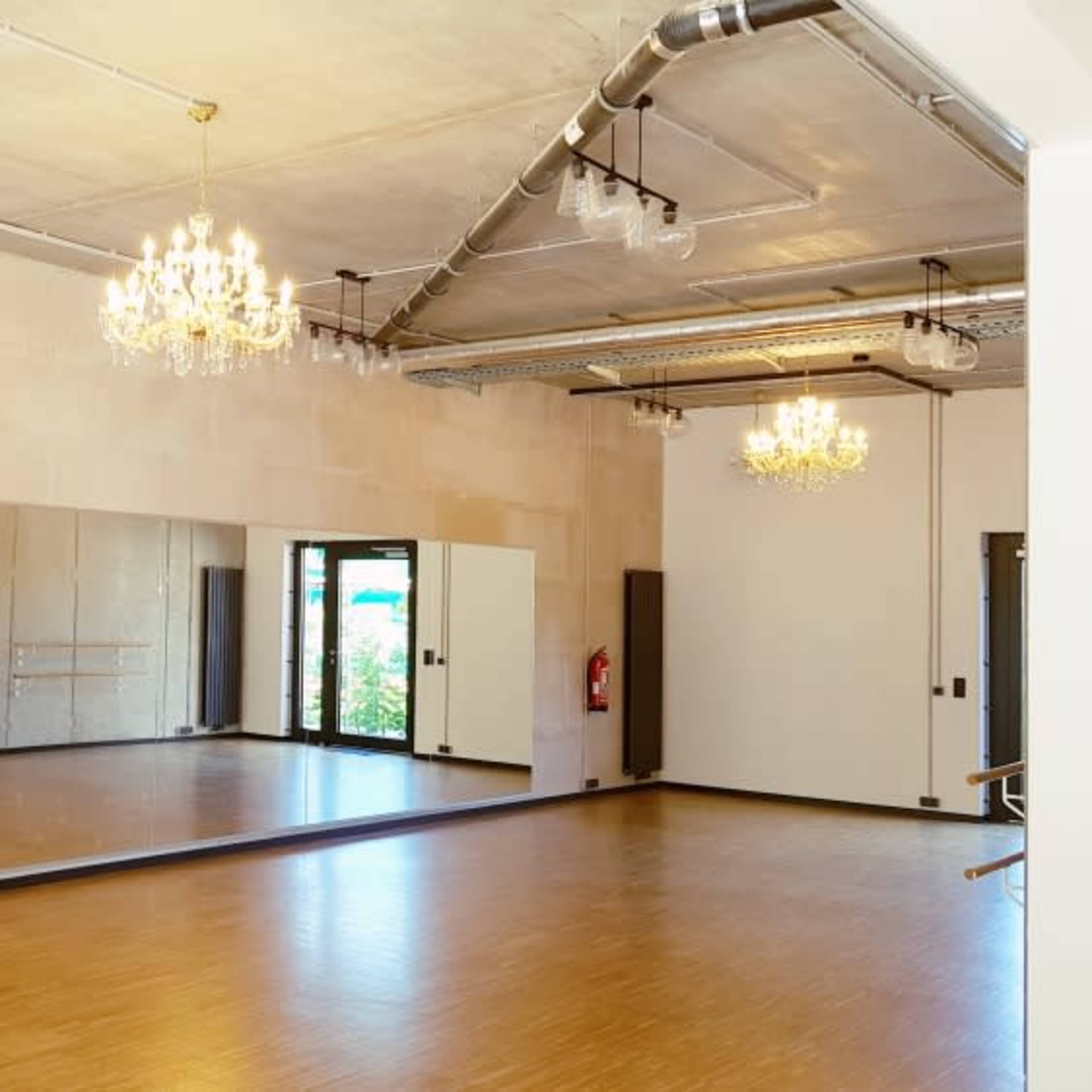 A bright, spacious dance studio featuring wooden flooring, large mirrors, and elegant chandeliers.
