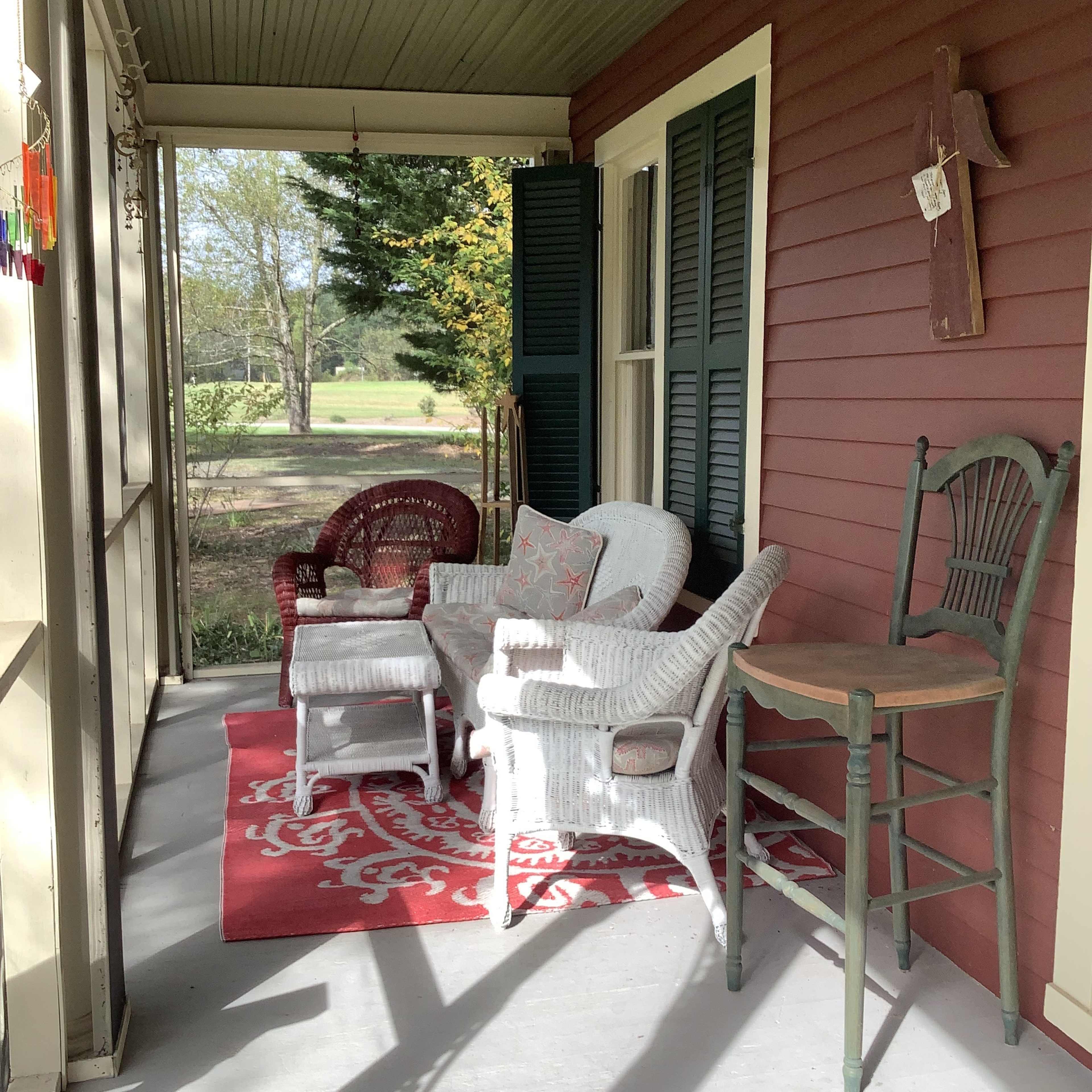 A porch features various seating options, including wicker chairs, a small table, and a patterned rug, with green foliage visible in the background.