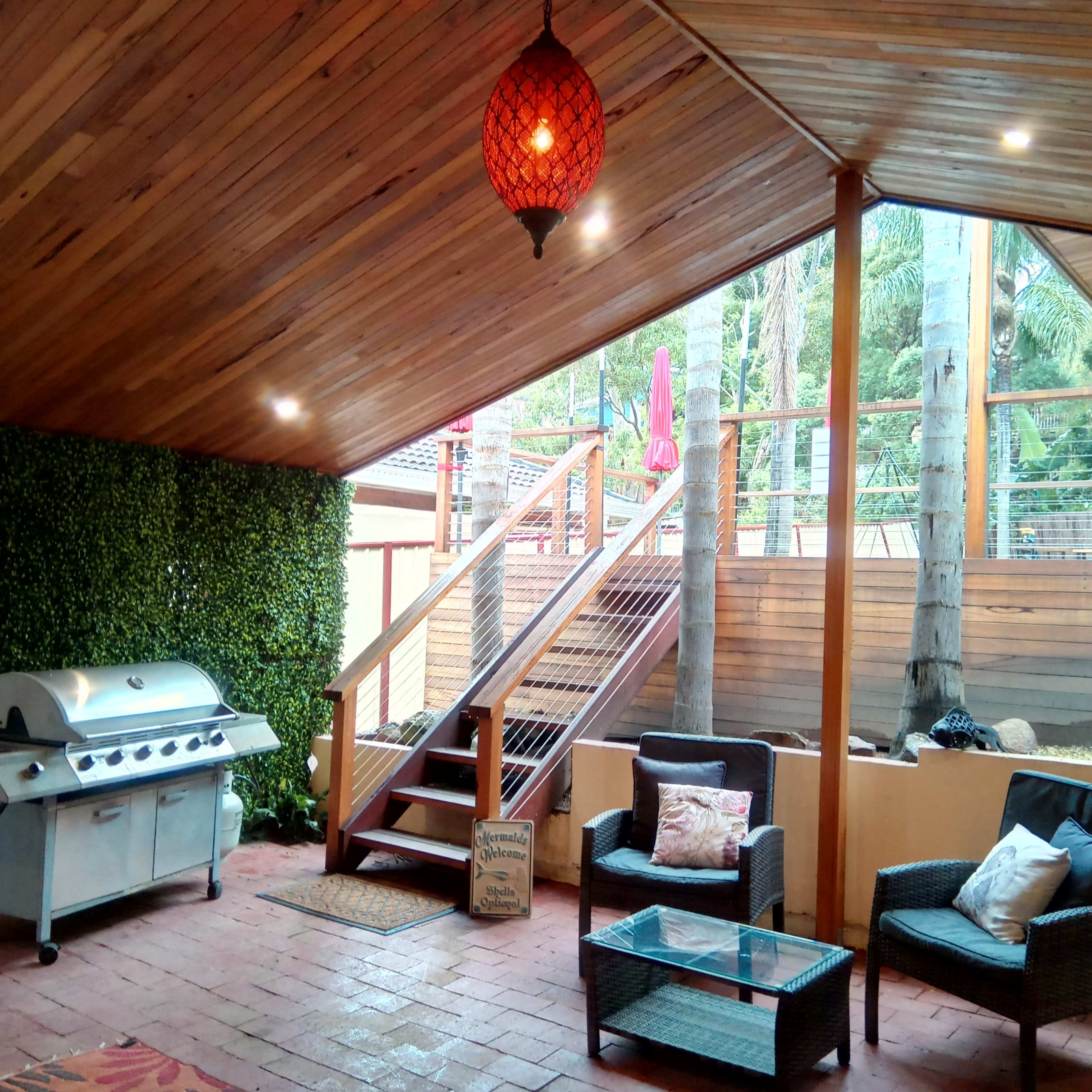 An outdoor patio features a grill, a glass coffee table, two chairs, and wooden stairs leading to a higher deck, all under a wooden ceiling with a hanging light.