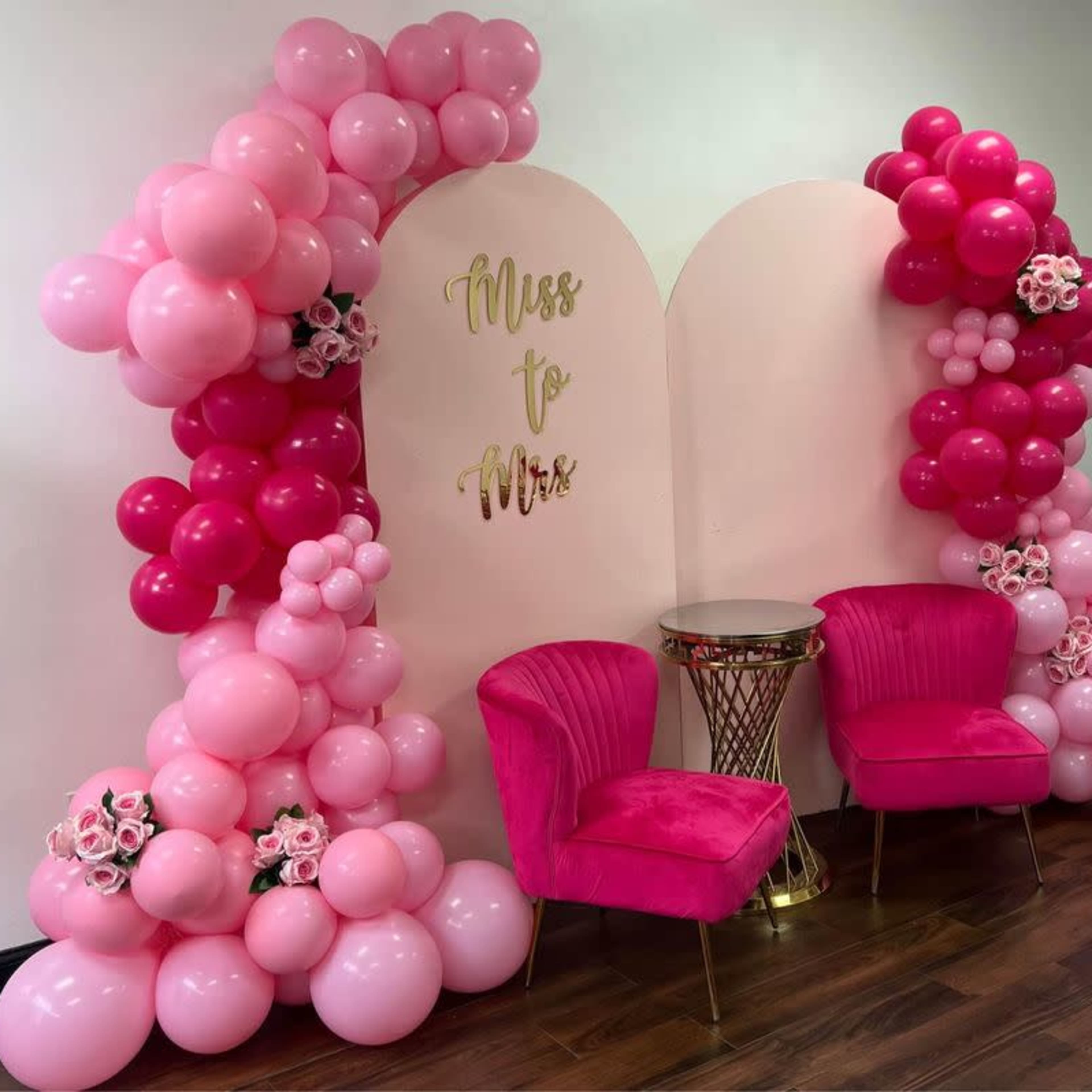 A decorative backdrop with pink and fuchsia balloons arranged around the words "Miss to Mrs," flanked by two pink velvet chairs and a small gold table.