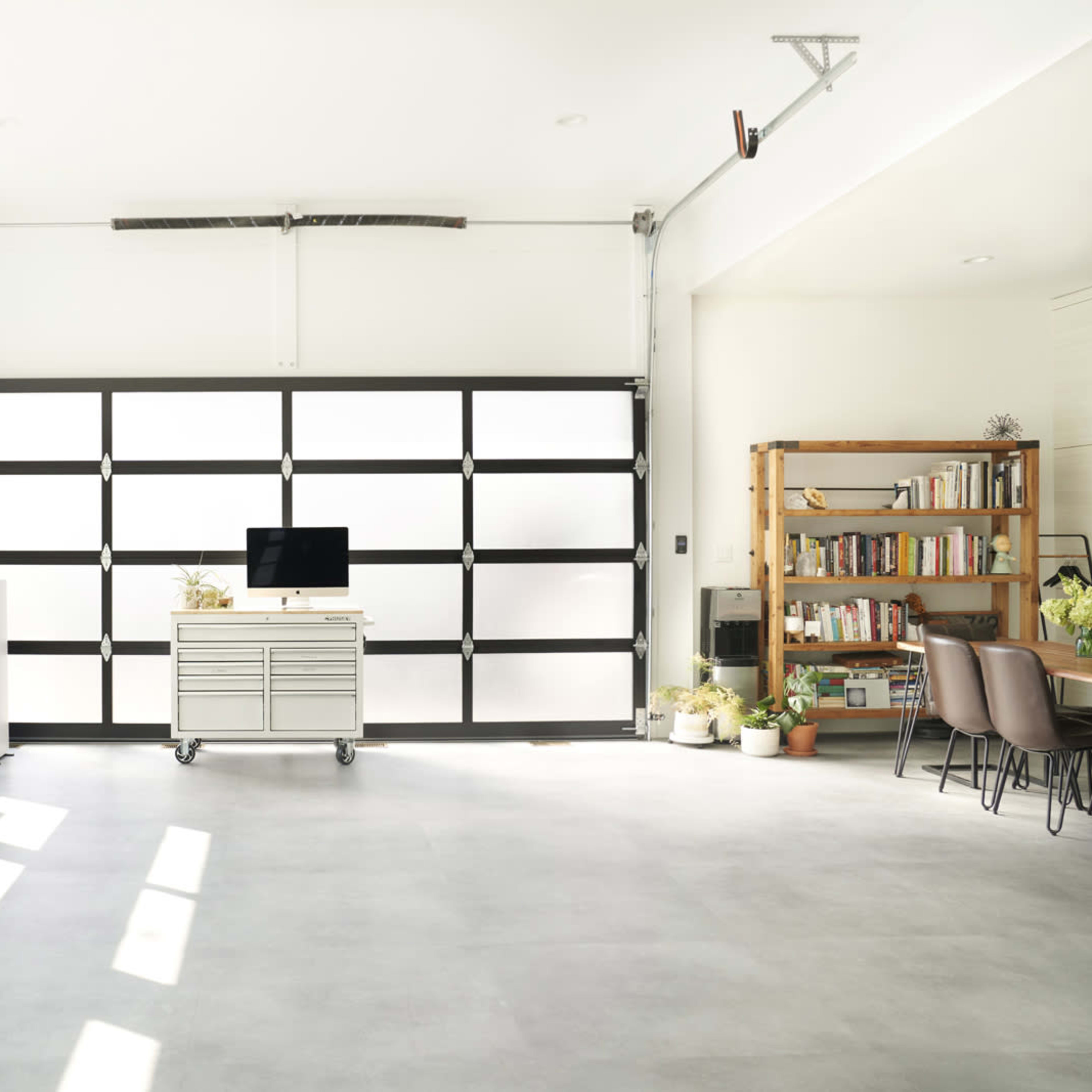 A modern garage with a large glass door, a workbench on wheels, a television, a bookshelf filled with books, and potted plants.