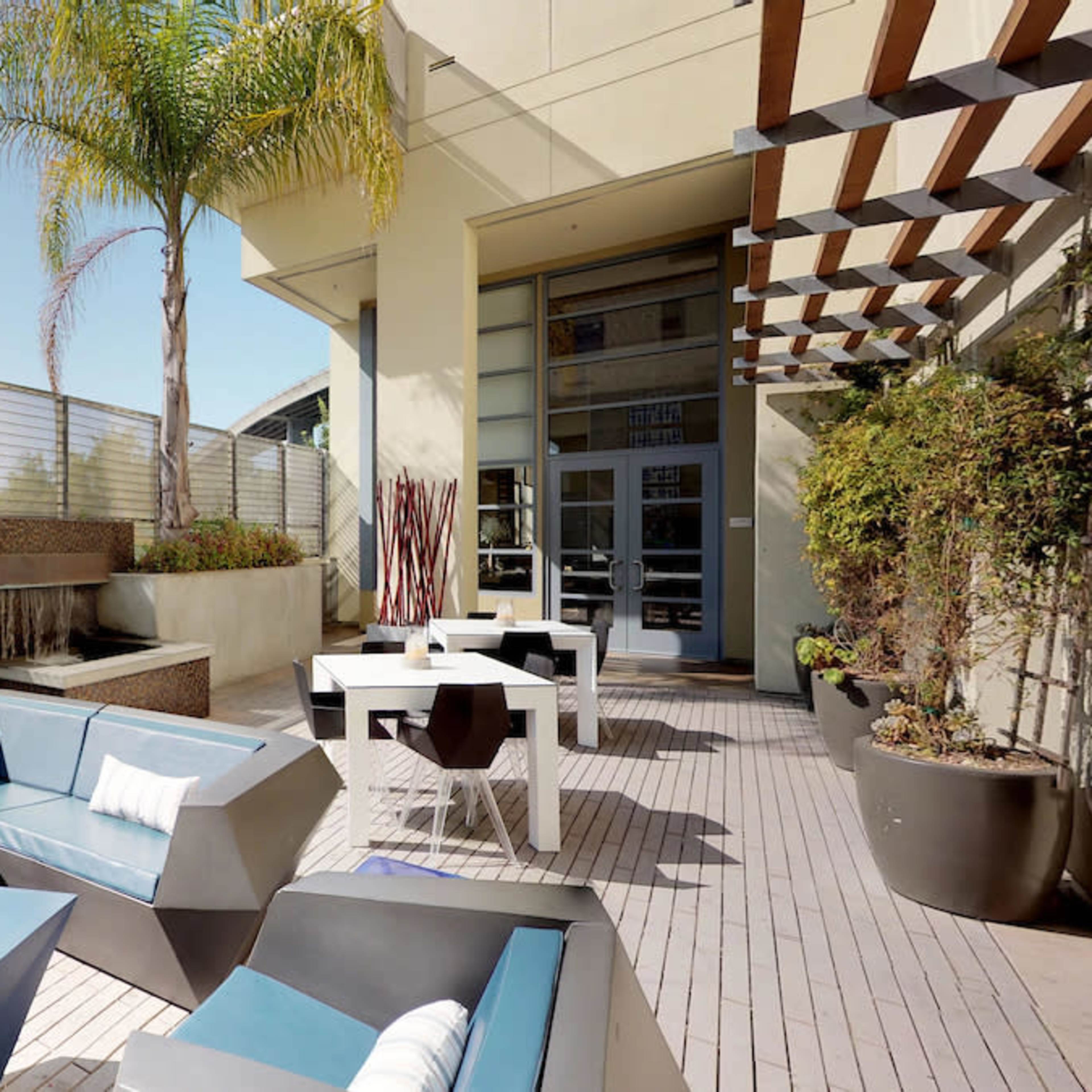 A modern outdoor patio area featuring a large sectional sofa, tables, and decorative plants, with a water feature in the background.
